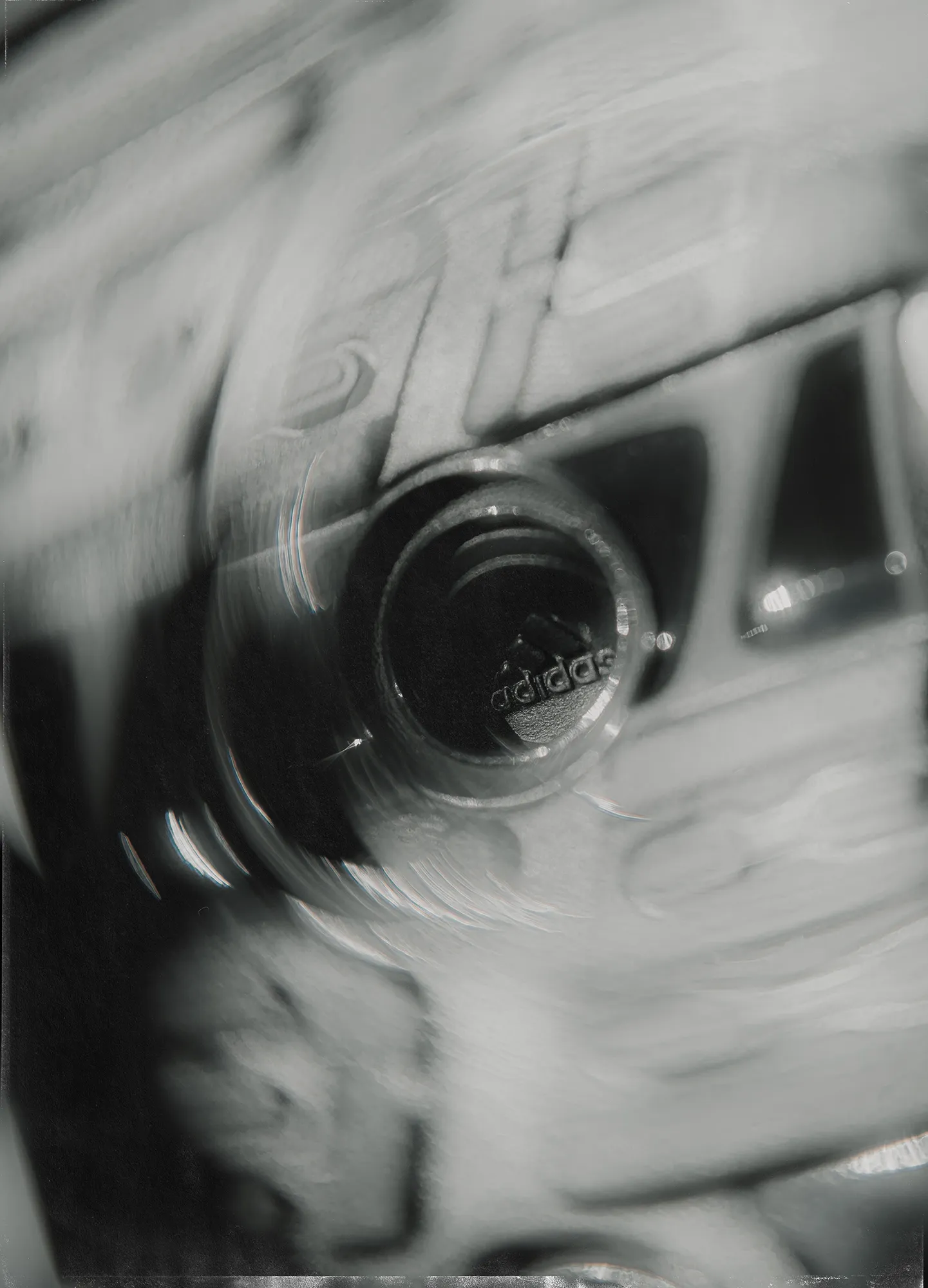 Black-and-white image of an Adidas sneaker floating against a dark background, surrounded by circular motion blur, with a textured, film-like border that emphasizes speed and energy.