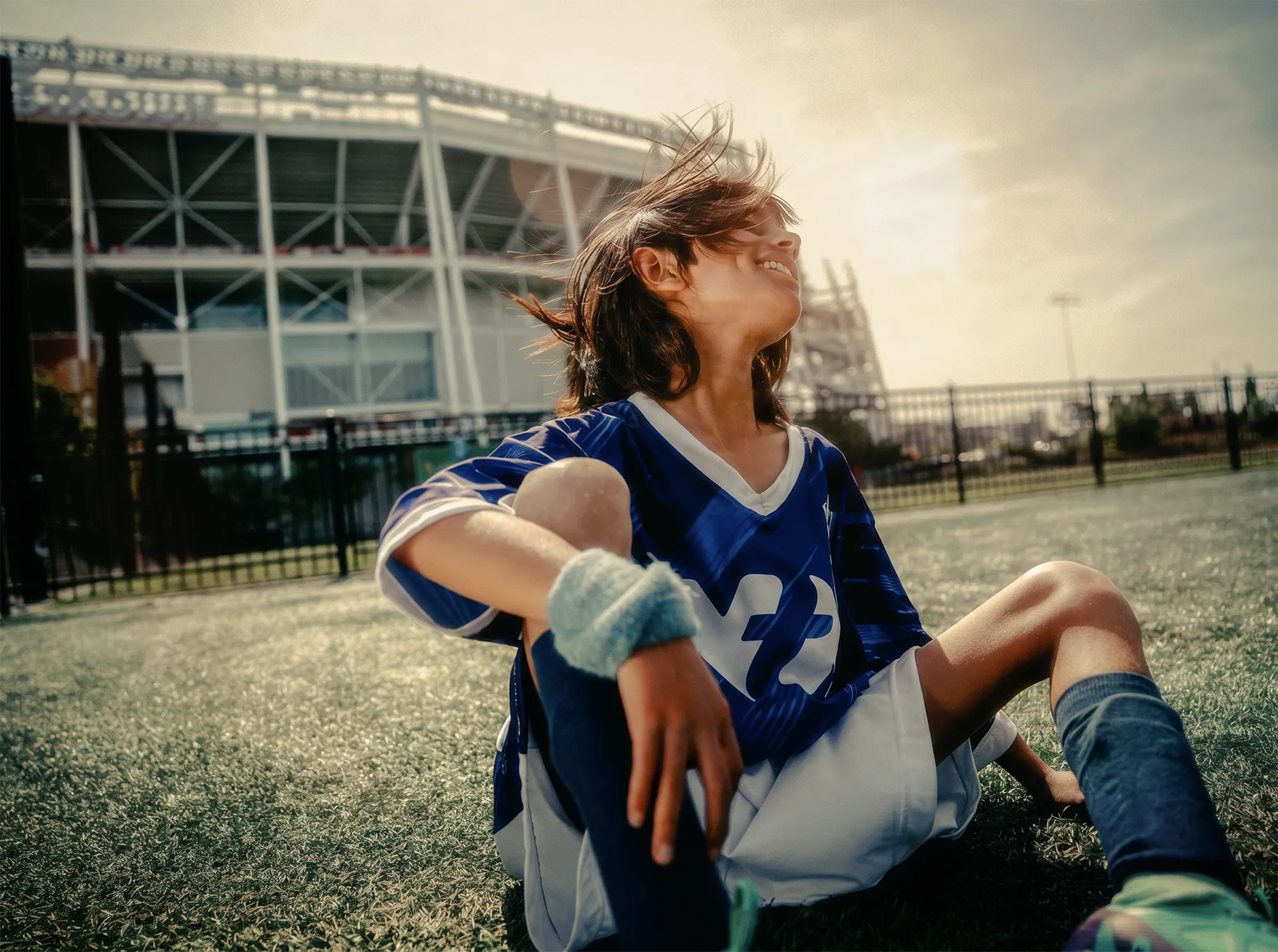 A smiling young soccer player in a blue jersey sits on a sunlit field in front of Levi's stadium in North California, with wind blowing through their hair.