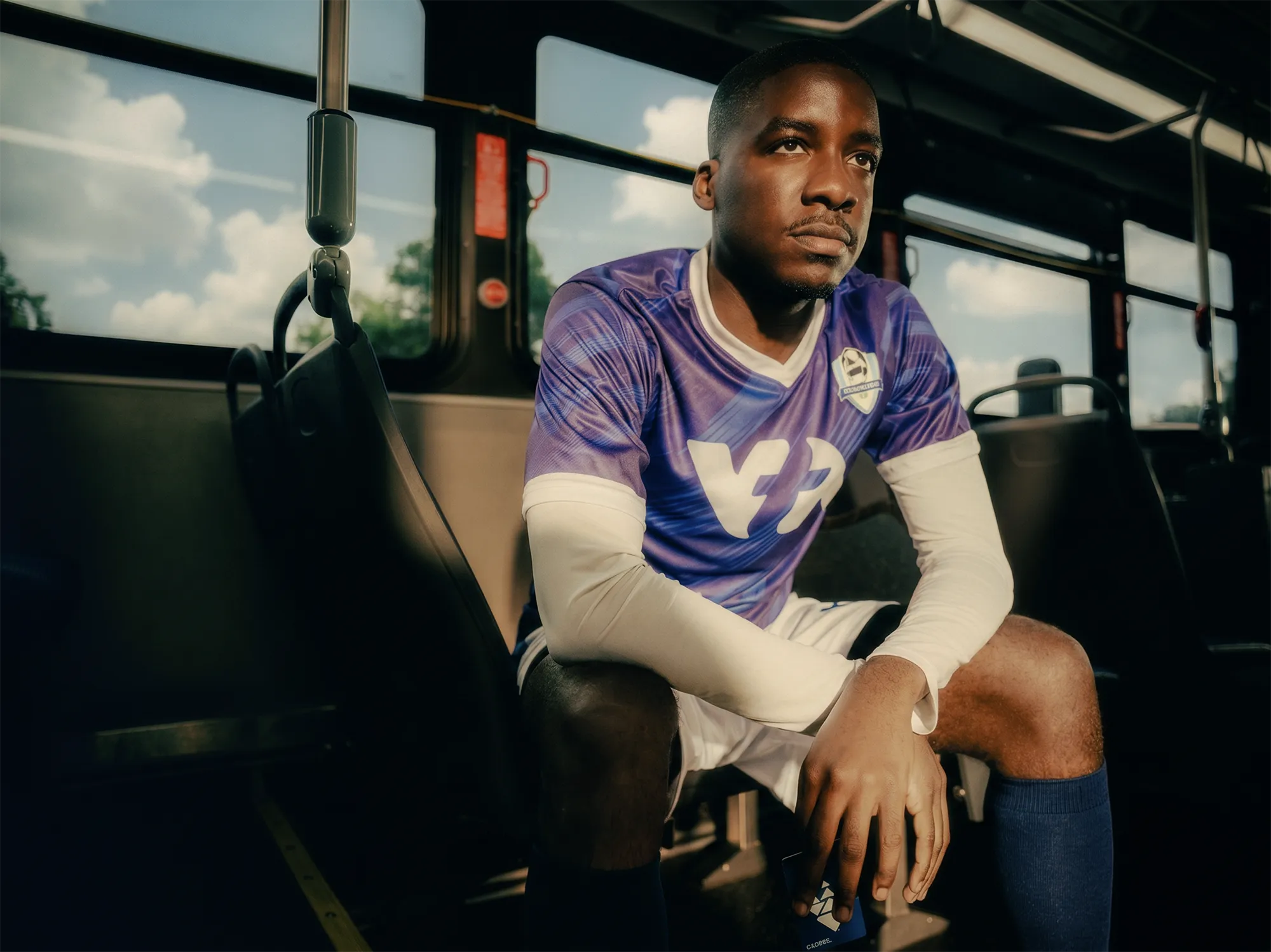 A soccer player in a blue jersey sits on a VTA city bus in North California, looking thoughtfully out the window while traveling to a game, holding a Clipper card.
