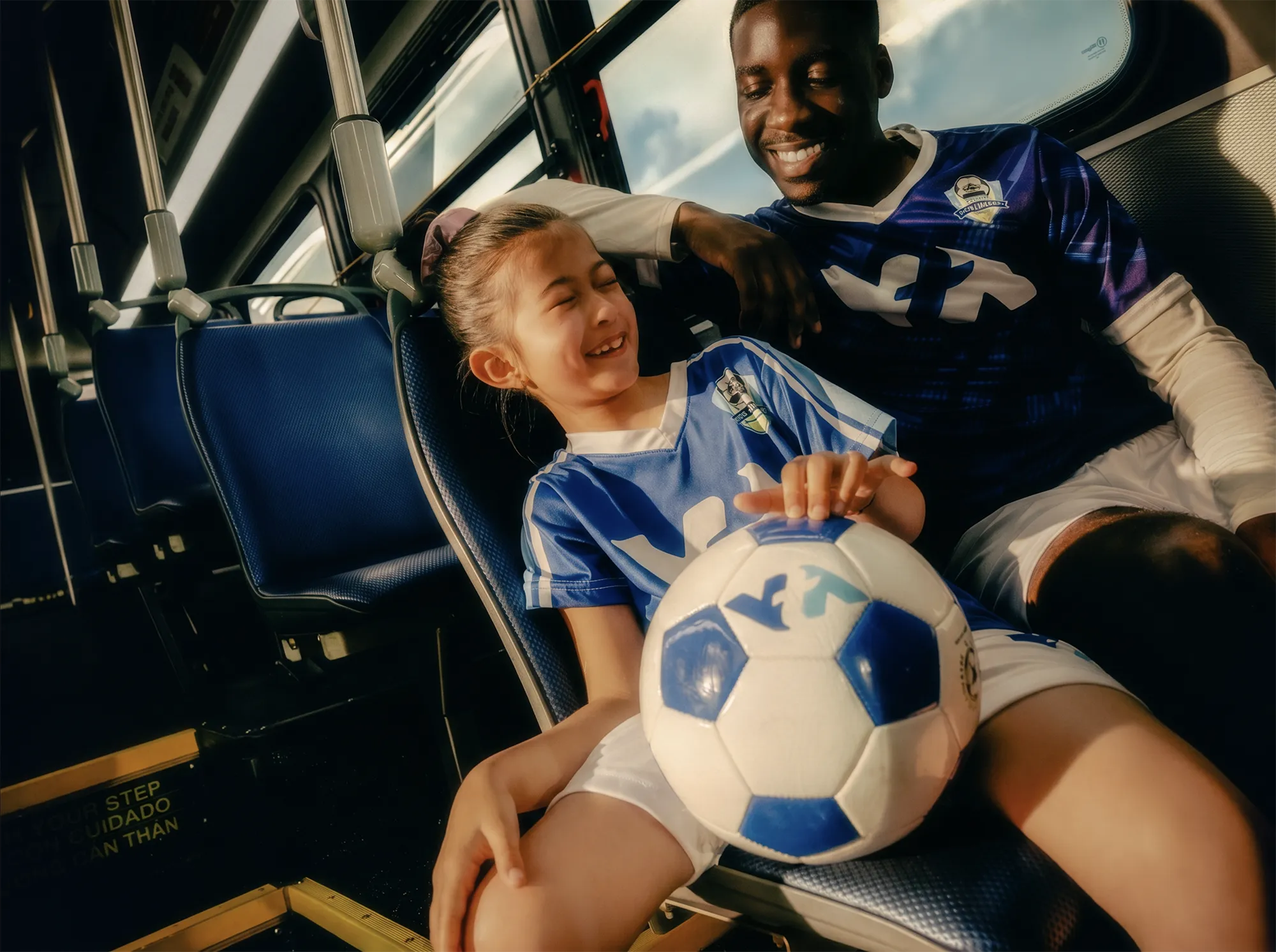 A young soccer player and an adult teammate sit together on a city bus in blue uniforms, smiling and chatting while holding a soccer ball during the ride to a match.