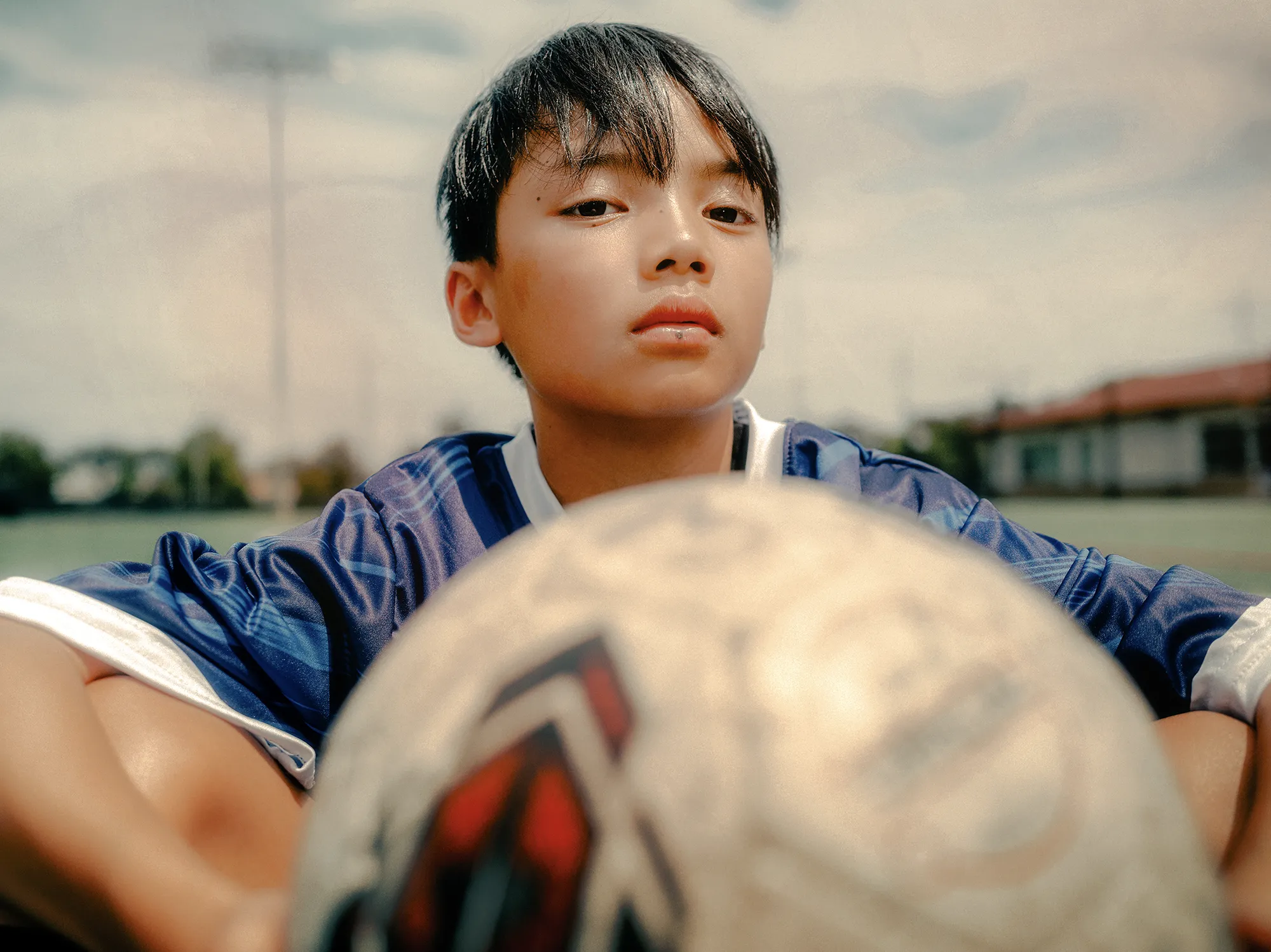 A young soccer player in a blue jersey sits on a field with a soccer ball in the foreground, looking confidently toward the camera.