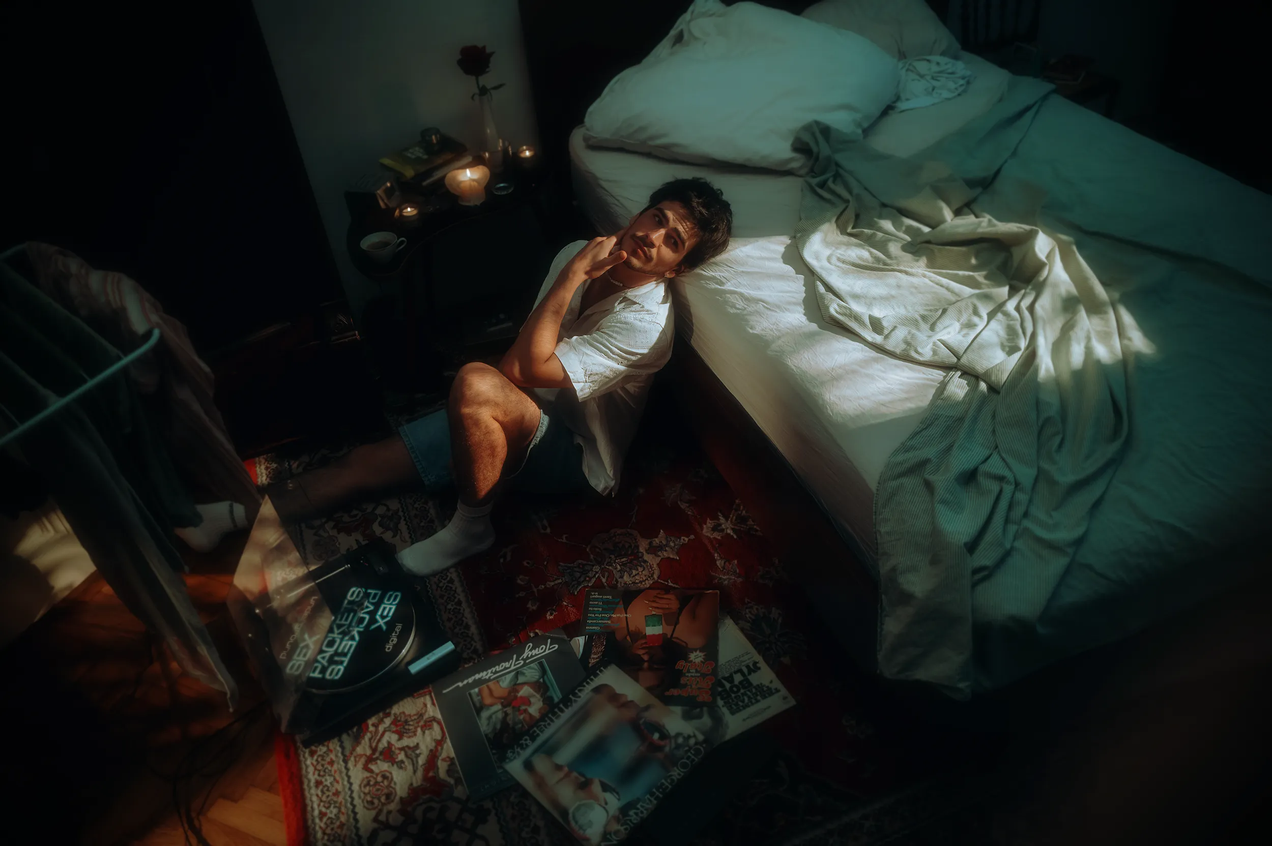 Unit still photography and advertising photography of a young man sits on the floor beside an unmade bed, surrounded by candles and vinyl discs in a dimly lit bedroom with a beautiful afternoon light coming through the window.
