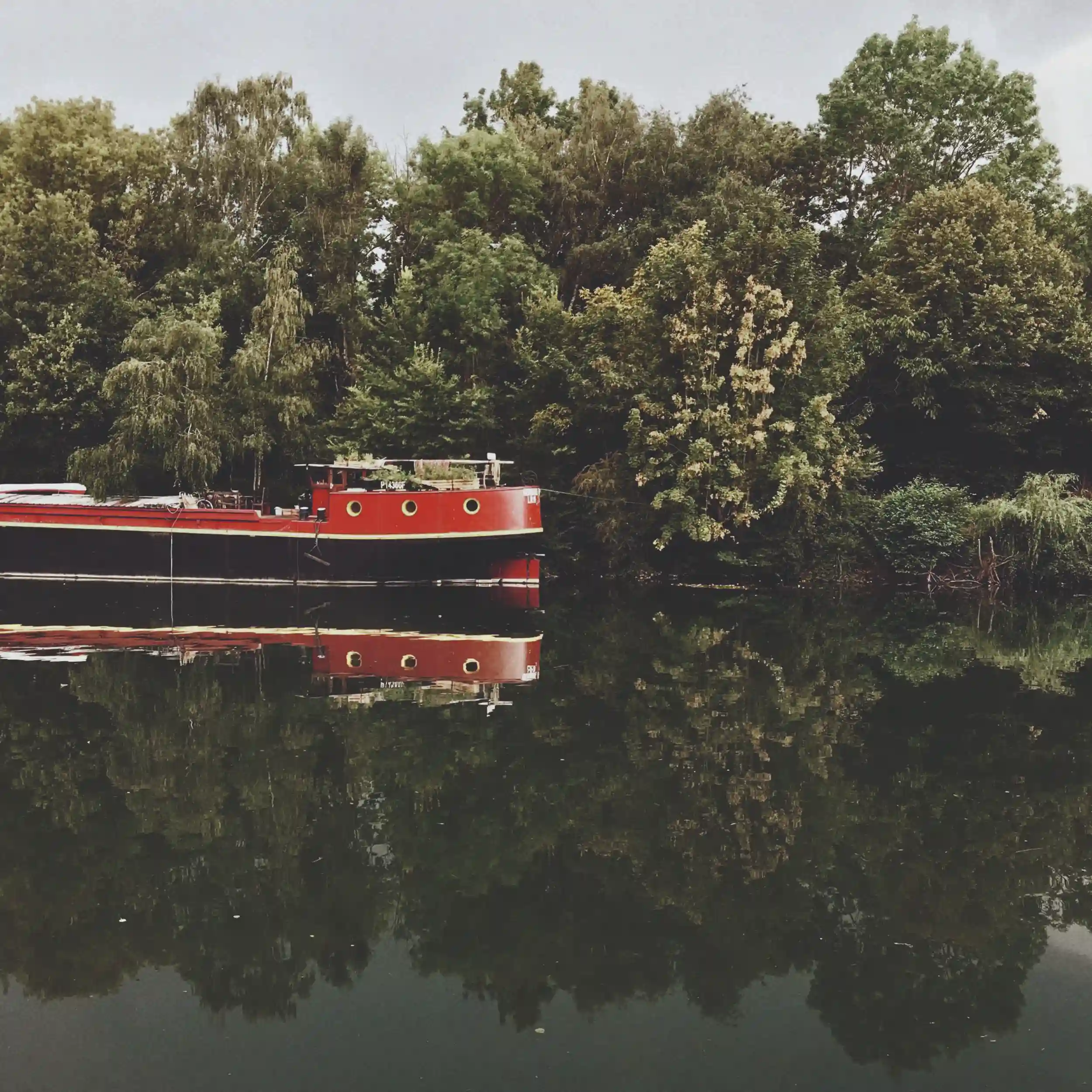 Bateau rouge amarré sur un cours d'eau calme avec des arbres verts reflétés dans l'eau.