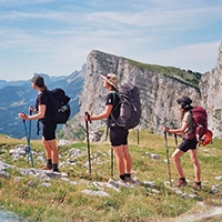 Trois randonneurs avec sacs à dos marchant sur un sentier de montagne herbeux avec un ciel bleu clair et des falaises en arrière-plan.