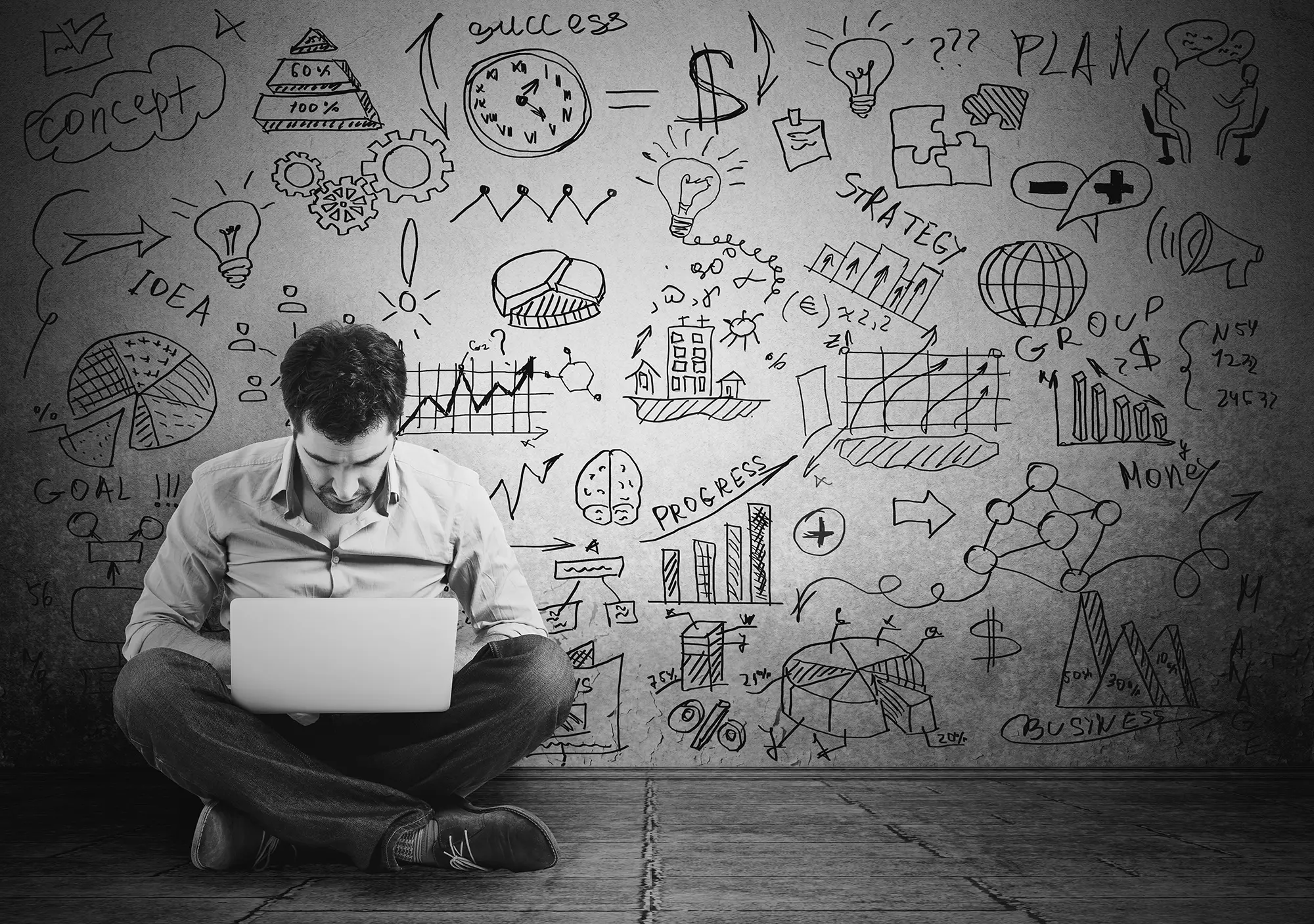 A man sitting cross-legged on the floor with a laptop in front of a wall covered in hand-drawn business doodles — ideas, strategy, charts and goals — in black and white