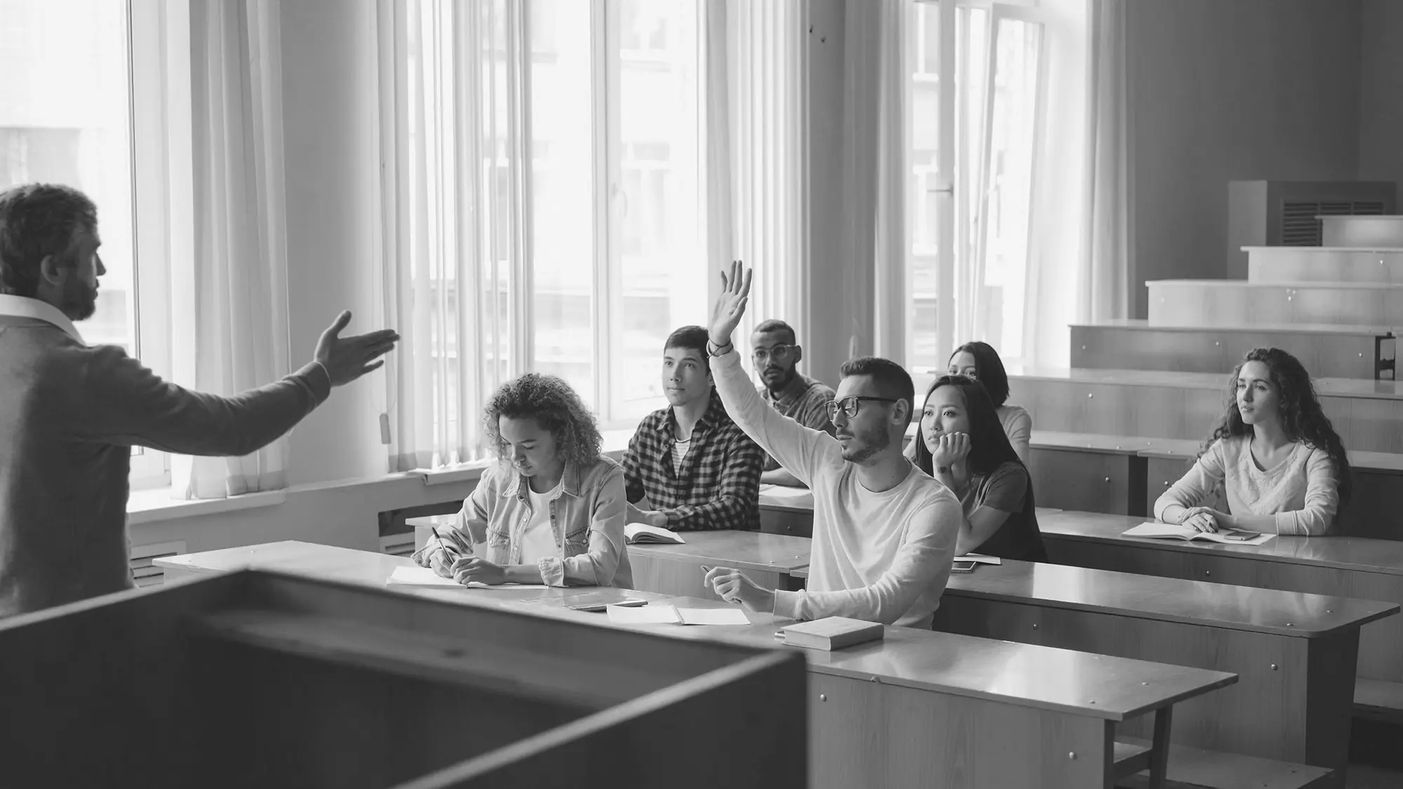 A lecturer gesturing to students in a classroom, one student raising a hand, black and white