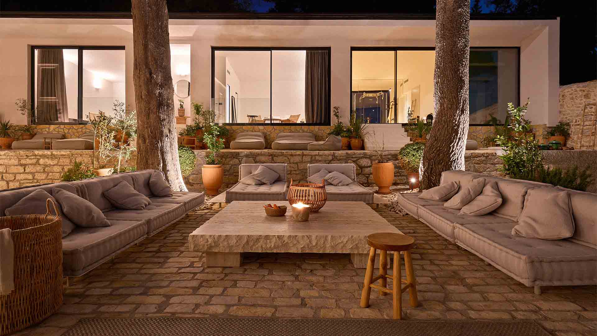Soft, atmospheric outdoor lighting illuminating a tranquil lounge space framed by large trees, with multiple rooms visible in the background.