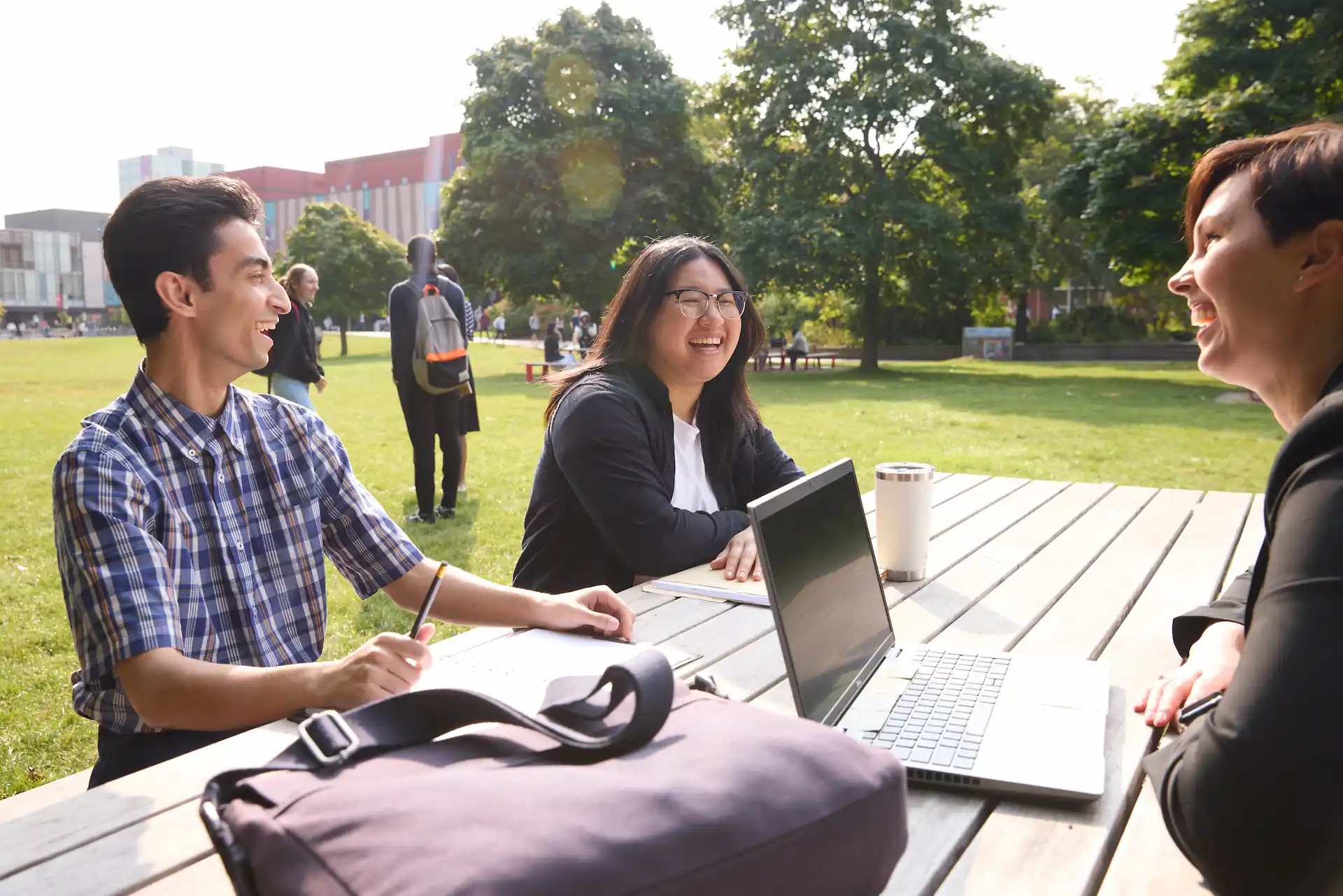 Three York University students chatting on a picnic bench while using AppsAnywhere on a laptop