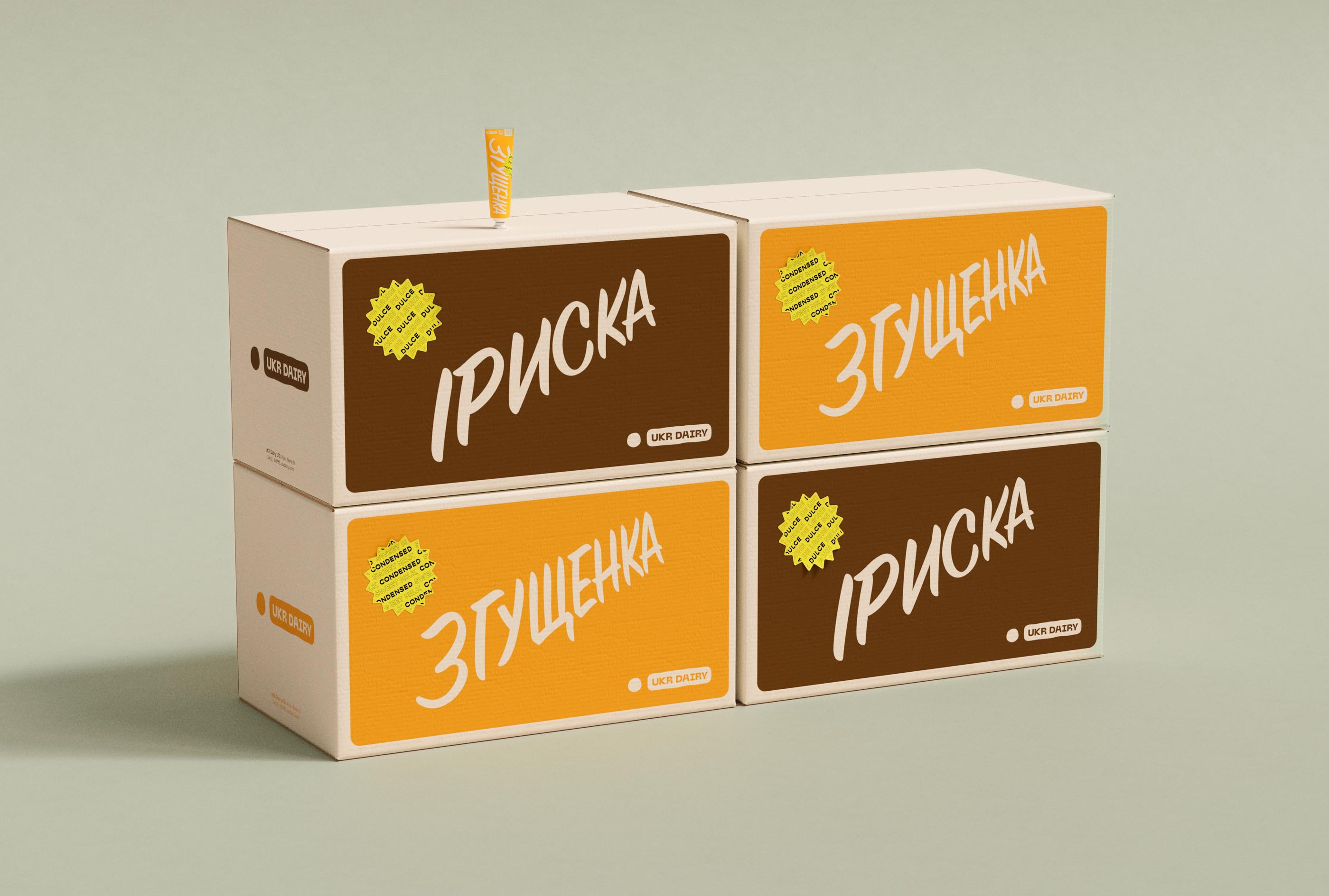  neatly stacked arrangement of UKR Dairy's condensed milk and dulce de leche boxes, showcasing the packaging design by a creative design agency. The image features two brown boxes labeled "ИРИСКА" and two yellow boxes labeled "ЗГУЩЕНКА" in bold white text. Each box includes a starburst label highlighting the product content. The minimalist and clean design enhances brand visibility and reinforces the quality of the products. A single tube of condensed milk is placed atop the stack, emphasizing the innovative and attractive packaging.