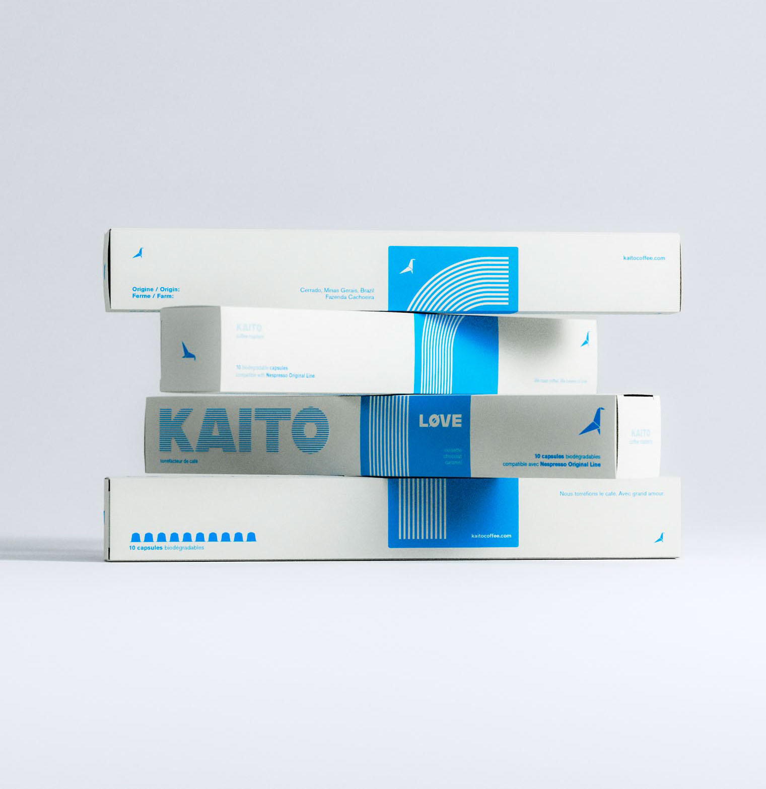 A visually engaging stack of four KAITO coffee capsule boxes arranged in an offset composition, emphasizing the modular and graphic nature of the packaging system. Each box features crisp white surfaces with a consistent electric blue accent, incorporating geometric shapes, linear patterns, and structured typography. The bold typographic logo contrasts with fine-line illustrative elements, suggesting a refined balance between graphic design clarity and expressive branding. Ideal lighting and a soft backdrop highlight the thoughtful art direction and brand identity created by a packaging design studio specializing in sustainable coffee products.