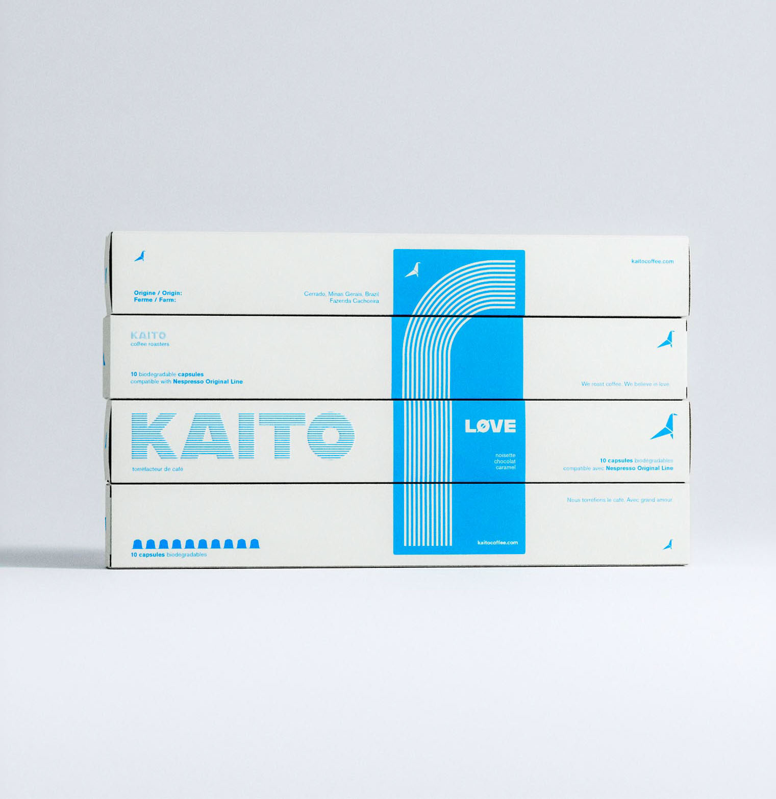 A neat vertical stack of four coffee capsule boxes, designed in a minimalist cream-white and cobalt blue palette, set against a neutral grey background. The packaging design features bold sans-serif typography, a stylized linear arch motif, and clean typographic hierarchy. The brand name "KAITO" is prominently displayed using striped lettering, while the word "LOVE" appears in contrasting bold white on a blue field. The layout integrates bilingual text and includes capsule compatibility information in small print. This image exemplifies considered packaging design, modern branding, and structured graphic design work from a packaging design agency.