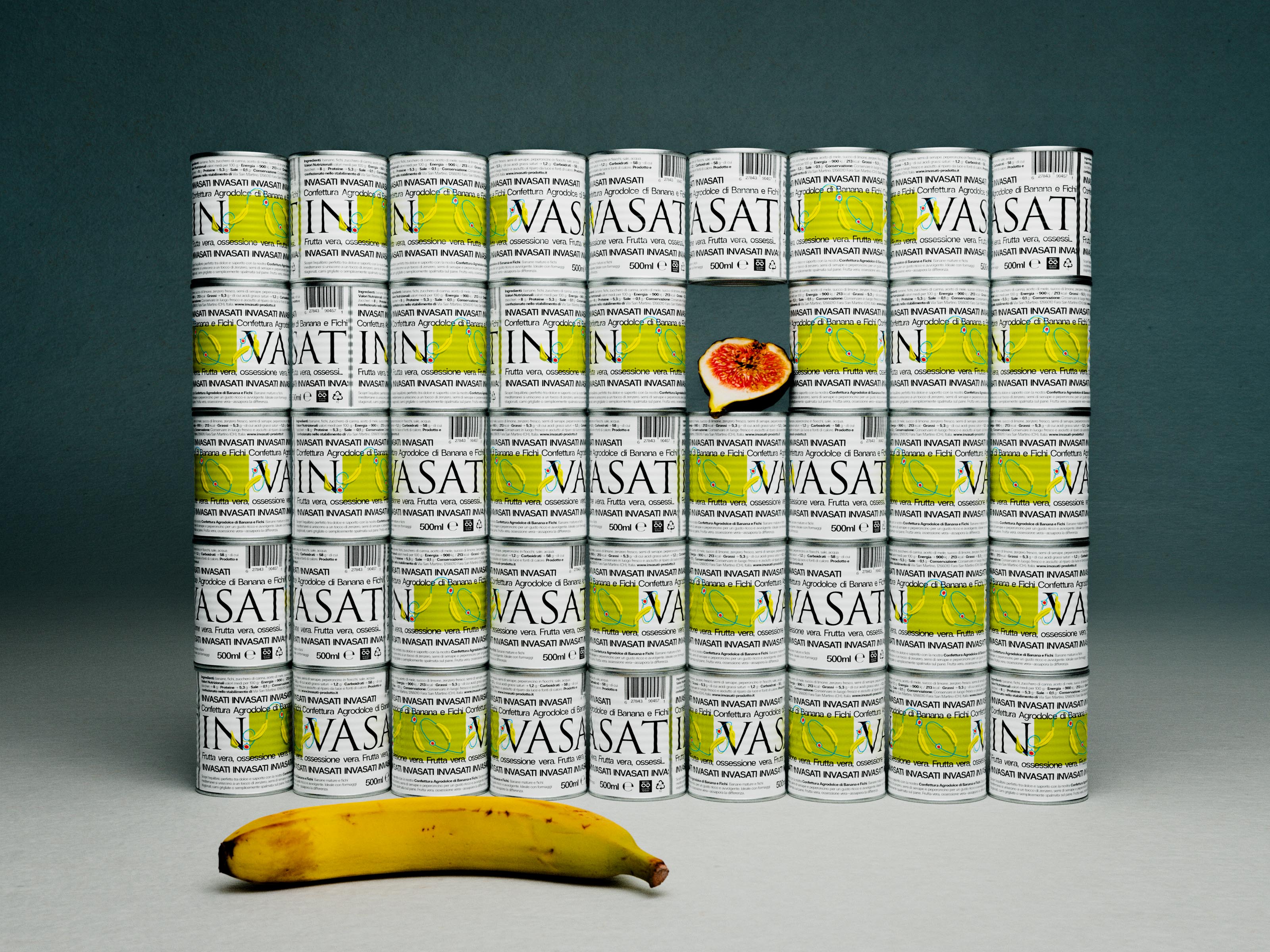 The photograph presents a meticulously ordered wall of slim aluminium cans, each wrapped in a crisp white label dominated by elegant black serif typography spelling “VASATI”. A chartreuse block illustration of a segmented banana slices horizontally across every label, injecting energy and creating a repeating rhythm that underscores the packaging design agency’s emphasis on clarity and shelf impact. The cans are stacked in precise columns on a cool grey surface, their collective pattern becoming a graphic-design grid that signals disciplined art direction. Mid-centre, a ripe fig is wedged between rows, its ruby seeds and ochre flesh disrupting the uniformity and hinting at the fruit purée within—an identity design flourish that adds narrative without clutter. In the foreground a gently speckled banana arcs across the frame, grounding the scene with tactile authenticity that a branding studio might use to signal freshness. Diffuse overhead lighting produces soft, studio-grade shadows, ensuring every barcode, recycling icon and 500 ml detail remains legible while the deep teal backdrop recedes, keeping focus on the product. The composition exemplifies contemporary creative direction where minimal packaging design meets expressive storytelling, demonstrating how a design agency can balance precision with emotive cues to elevate everyday grocery items