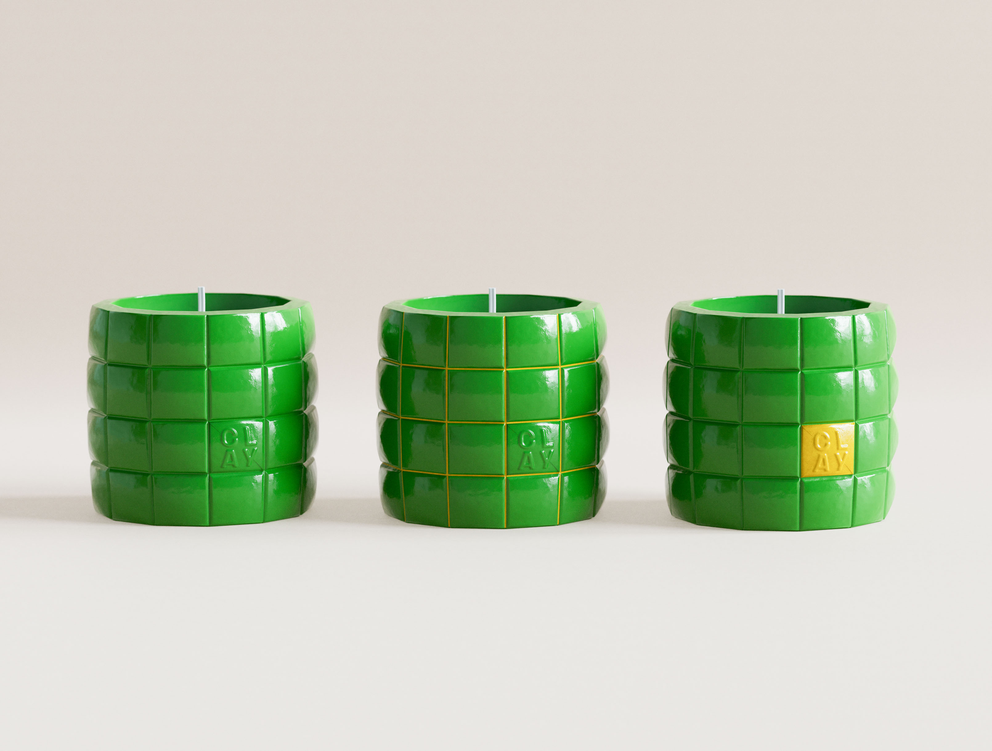 Three emerald green CLAY grid candles in studio lineup. Left: monochrome gloss green with embossed CLAY mark in tile cell. Centre: same form with yellow grid lines dividing puffy tile segments. Right: monochrome green with single yellow CLAY accent tile. Glossy ceramic-form surface, embossed logotype. Visual identity design system and structural packaging design for a New York candle brand by a graphic design studio.