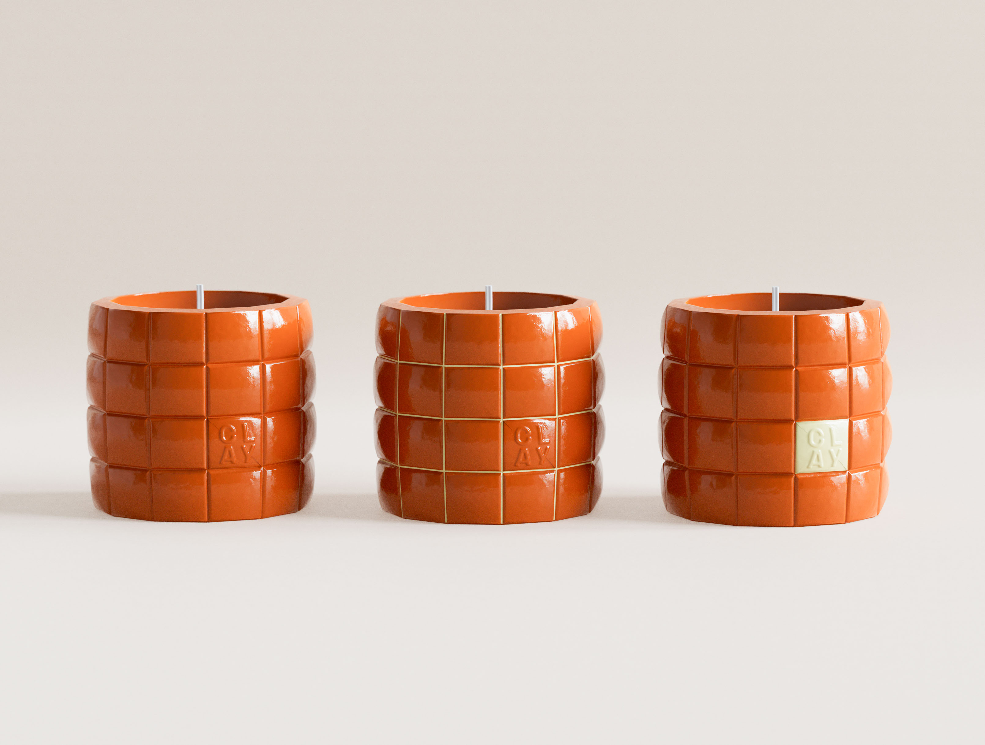 Three terracotta orange CLAY grid candles in studio lineup. Left: monochrome gloss orange, embossed CLAY mark. Centre: orange with olive-green grid lines. Right: orange with pale cream CLAY accent tile. Glossy ceramic-form glaze across all three. Color-variant visual identity design system and structural packaging design for a New York candle brand by a graphic design studio.