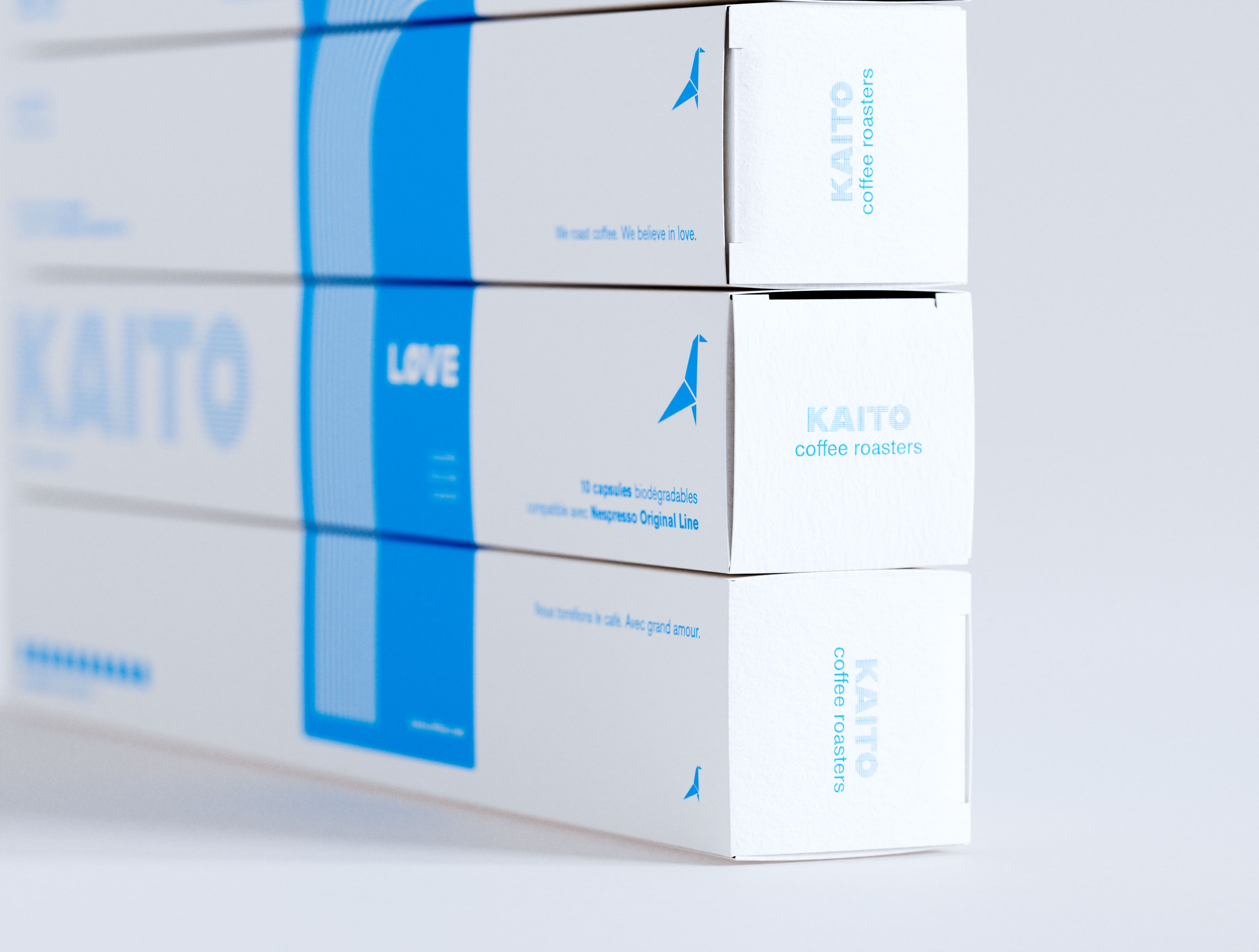 Close-up of the right end panels of KAITO coffee capsule boxes stacked four high. White coated paperboard, single-color cyan print. End panels read "KAITO coffee roasters" in clean geometric sans-serif with the origami bird logomark on each tier. Front face visible at left: "10 capsules biodégradables compatible avec Nespresso Original Line," "Nous torréfions le café. Avec grand amour." and LØVE variant on the blue band. Bilingual French/English label architecture and typography and graphic design by a packaging design studio.