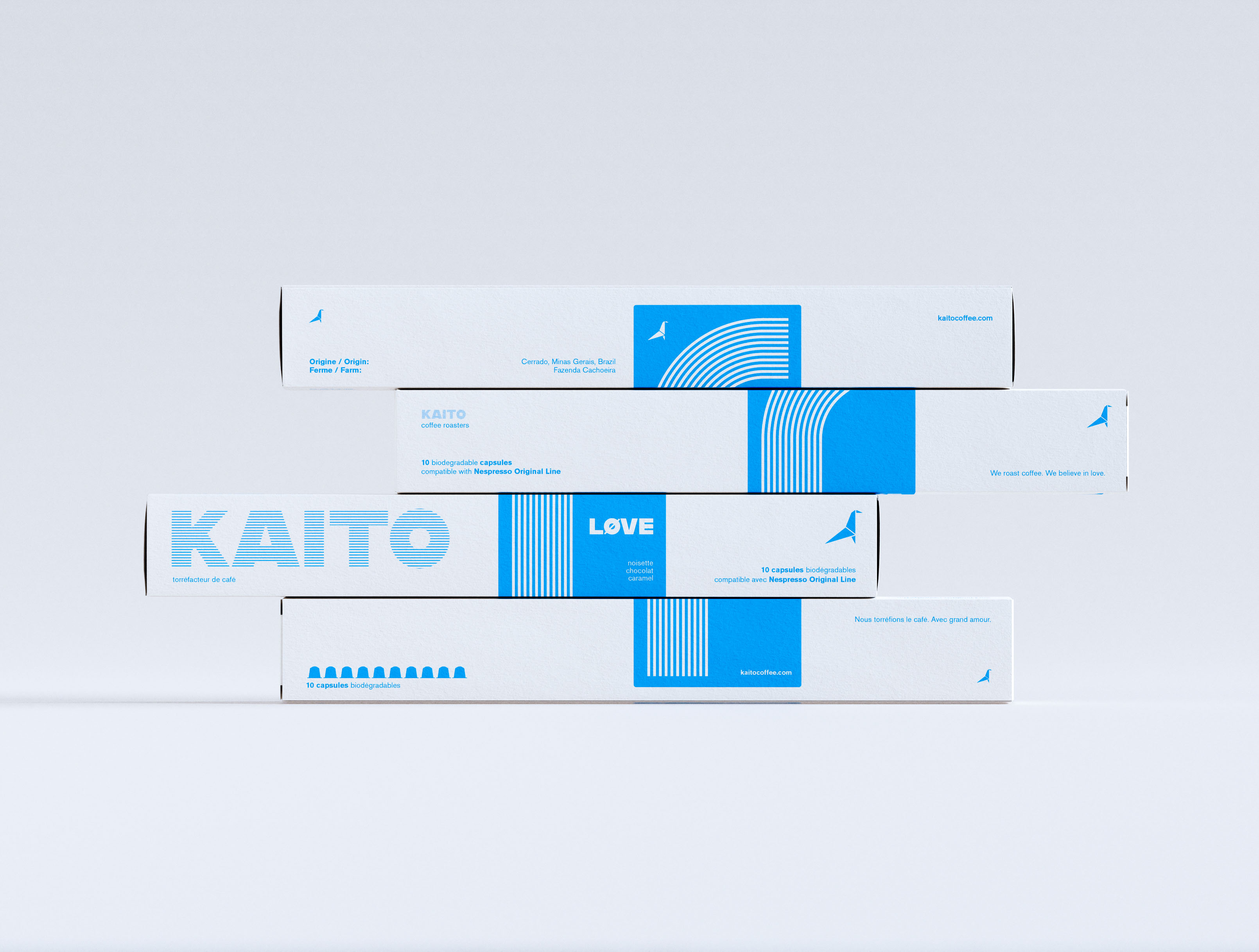 Four KAITO coffee capsule boxes in a horizontal-shift arrangement, each box displaced laterally to form a stepped profile against neutral grey. White coated paperboard, single-color cyan print. KAITO wordmark in striped type fill across the front face; LØVE variant on the blue vertical band with concentric arc stripe pattern. Bilingual French/English label system: "10 capsules biodégradables," "Nous torréfions le café. Avec grand amour." Origami bird logomark. Art direction and visual identity design by a packaging design studio.