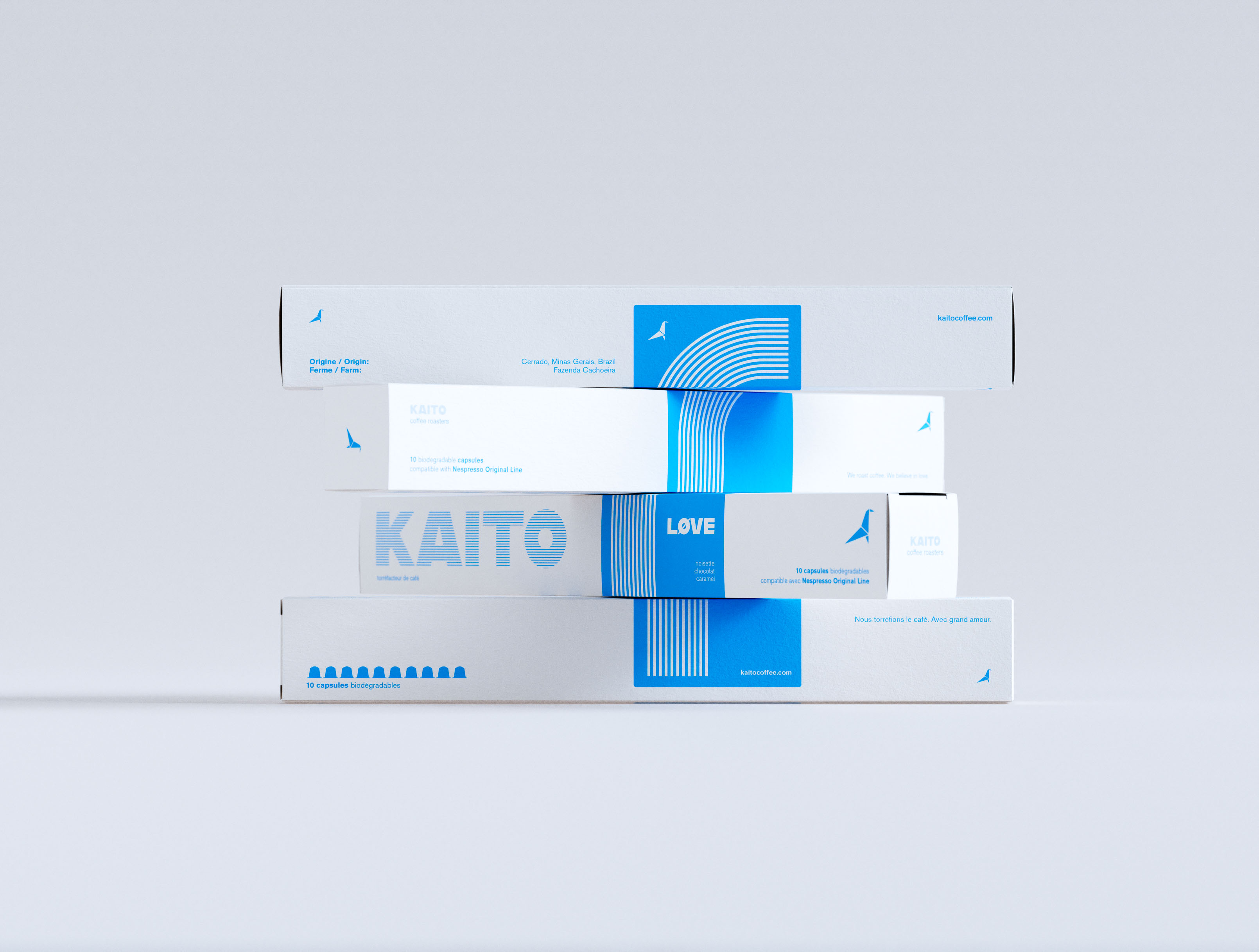 Four KAITO coffee capsule boxes in a cascading staggered arrangement, each box displaced progressively to the left on a neutral grey ground. White coated paperboard, single-color cyan print. KAITO wordmark in horizontal stripe type fill across the front face; LØVE variant wordmark and concentric arc stripe pattern on the blue vertical band. Bilingual English/French copy: "10 capsules biodégradables." Origami bird logomark on all boxes. Art-directed multi-format range composition — visual identity design system and art direction by a packaging design studio.