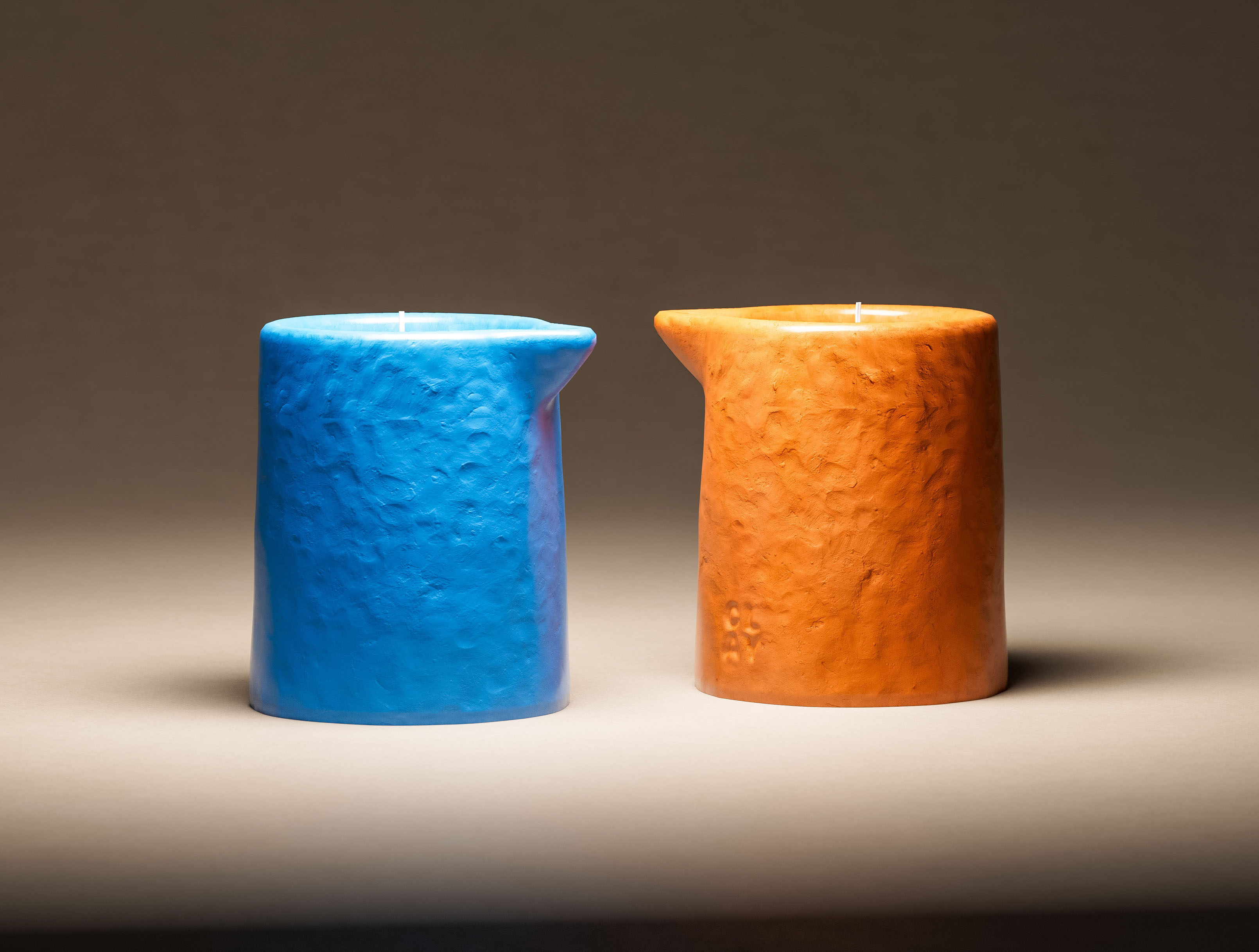Two CLAY pitcher candles in contrasting colorways — cobalt blue and terracotta amber — displayed side by side. Both in the brand's signature pitcher form with angled pour spout and rough hand-textured ceramic surface. CLAY embossed logotype on each vessel. Visual identity design system — a signature shape deployed across a multi-colorway range as a coherent brand identity, by a packaging design studio in New York.