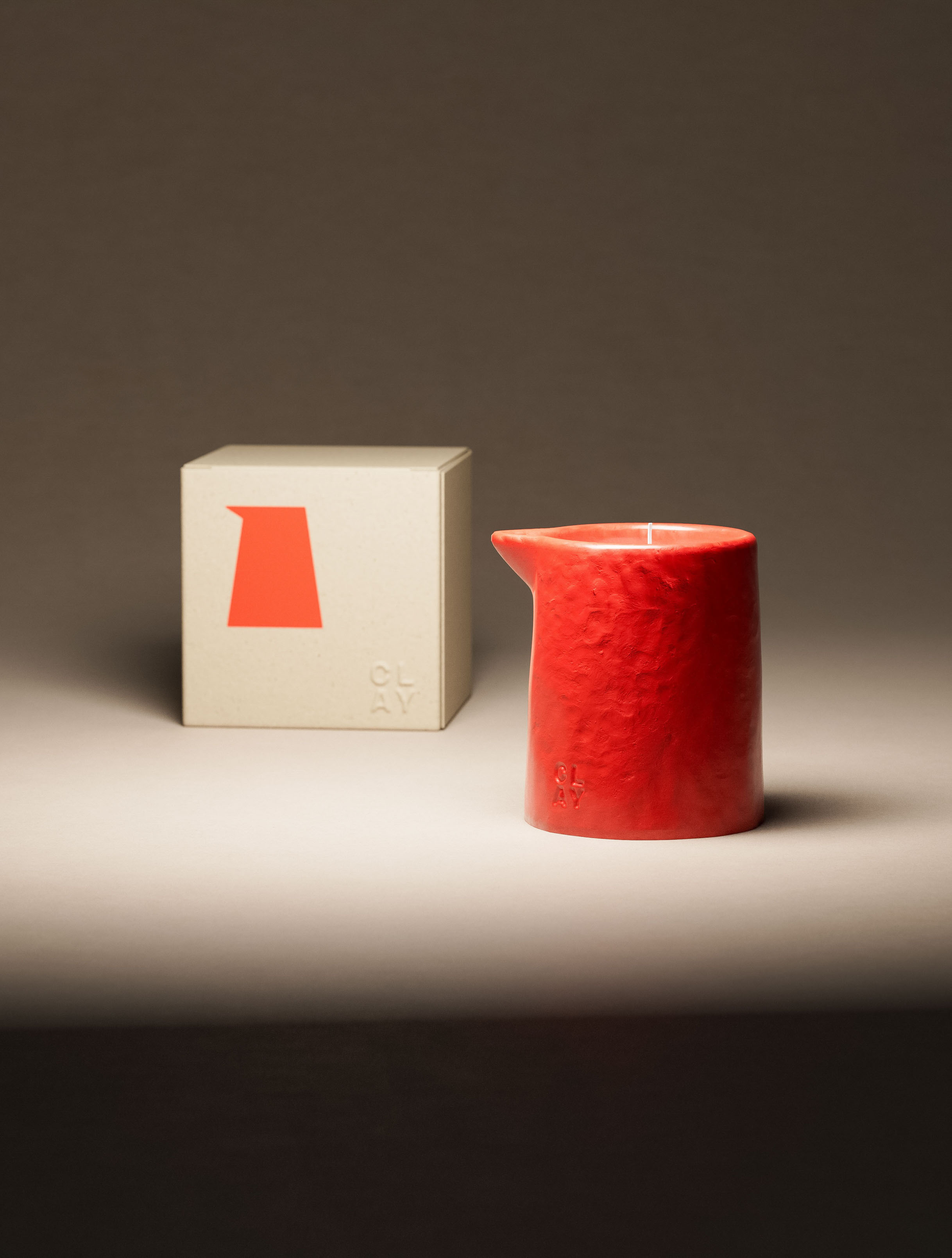 CLAY pitcher candle in coral-red glaze beside its natural kraft cube box, portrait crop. Signature pitcher vessel with angled pour spout and rough hand-textured clay surface. Orange pitcher icon and CLAY stacked wordmark on the box front. Embossed CLAY mark on the ceramic body. Brand identity and packaging design — the vessel silhouette used as the brand mark mirrors the physical product form, by a branding studio in New York.