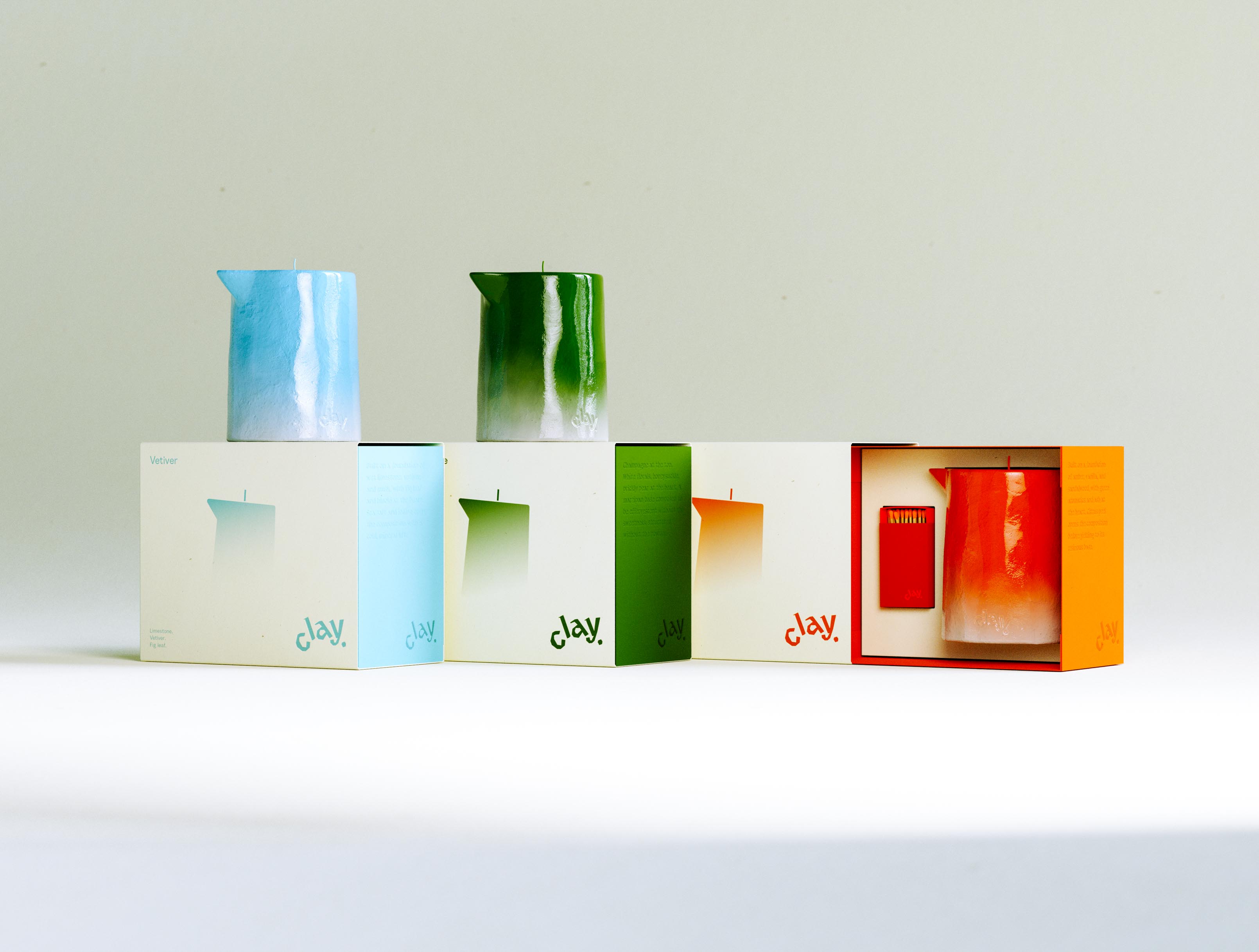 A wide line-up of the complete CLAY range: three cream cartons run left to right showing fronts and scent-coloured side panels, the blue Vetiver and green Champagne candles perched on the first two boxes while the orange Amber box at right is opened to reveal its candle beside a colour-matched orange matchbox with matches in the drawer. The "Vetiver" front, gradient silhouettes and colour-coded "clay." wordmarks step across the row, tying printed system, sculptural object and gift architecture into one frame. Bringing a three-scent candle line to launch at once — every carton, vessel and bespoke matchbox produced to spec and to schedule — is the end-to-end packaging and production Art of Packaging runs for a new home-fragrance brand.