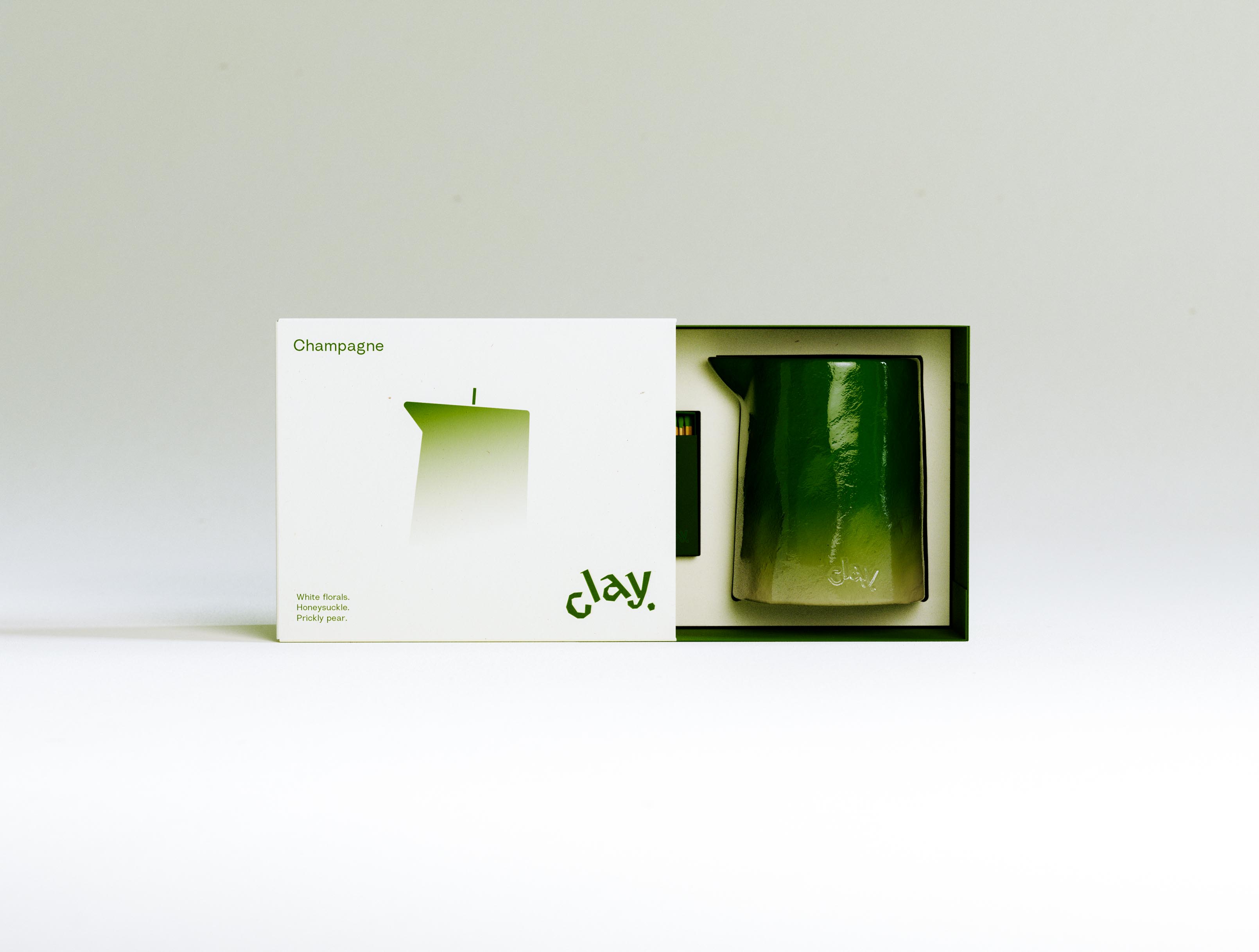 The CLAY Champagne gift box shown part-opened: the printed cream outer sleeve is drawn to the left to reveal a fitted deep-green tray cradling the glossy green pitcher-shaped candle, with a slim dark-green matchbox seated in its own recess alongside. The candle's lacquered surface catches a soft highlight and shows the lowercase "clay" deboss near its pale ombré base. The sleeve repeats the scent name, gradient silhouette, notes and olive wordmark; tray and candle share a single tonal green. It is a two-piece rigid sleeve-and-tray build in colour-matched board — the structural packaging and fitted-insert engineering a candle set needs to feel considered from the first slide of the box, with vessel, tray and bespoke matchbox all tuned to one palette.