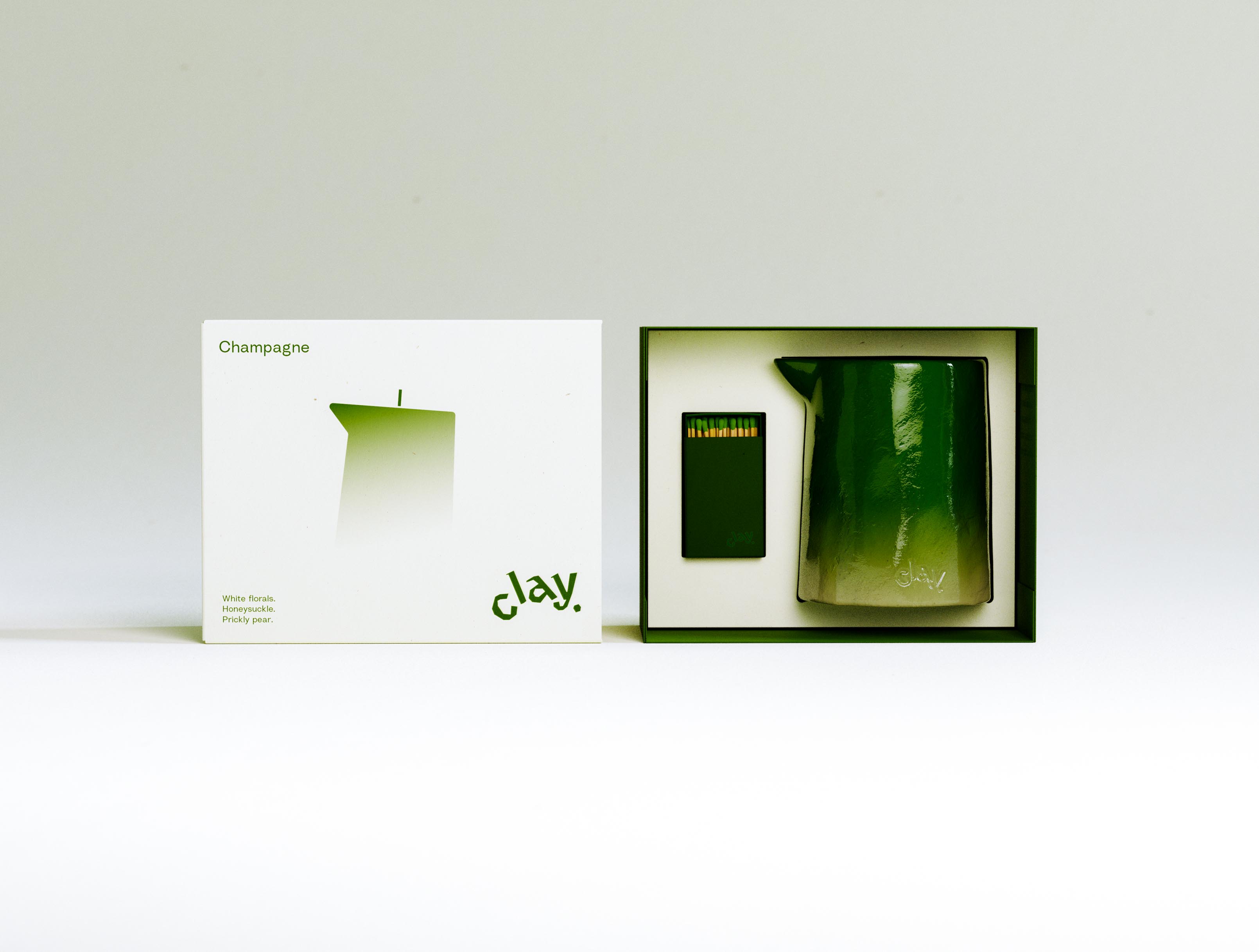 An open-presentation view of the CLAY Champagne set: the cream printed sleeve stands upright at the left while the deep-green tray at the right holds the glossy green pitcher candle beside an open dark-green matchbox, its green-tipped matches visible in the drawer. The candle's ombré runs from saturated green at the spout to a chalky pale base stamped with the lowercase "clay" deboss. Tray, candle and matchstick tips are tuned to one continuous green against the warm cream board. Details like colour-matched matches are where a launch earns its positioning, and producing them — sourcing the matchbox, specifying the tip colour, matching it to the wax — is part of how AOFP Studio carries a new candle brand from concept through to a finished, shelf-ready gift set.