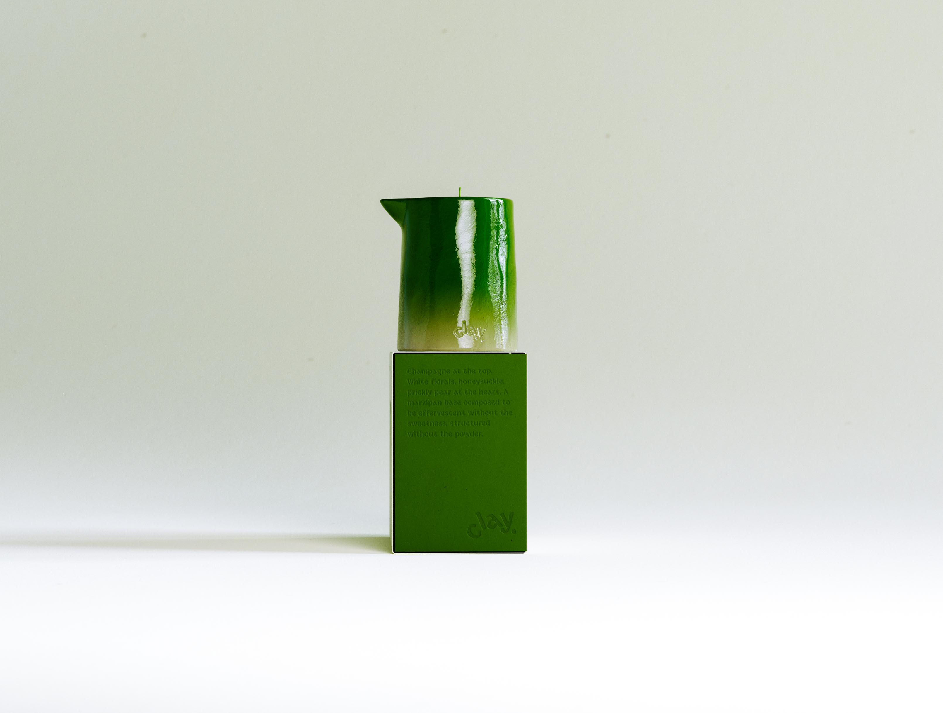 A pared-back vertical still life: the CLAY Champagne carton stood on end to present its saturated green side panel — embossed with the scent-pyramid copy and "clay." wordmark — with the glossy green candle balanced on top so green floods the frame against warm cream. The candle's lacquered ombré, pinched spout and single wick read crisply, while the blind-deboss copy catches a low raking light. Reduced to colour, form and finish, it exposes the logic under the range: one saturated hue owns each scent, carried identically across box, wax and wordmark. Building a colour-blocked, multi-SKU system this disciplined — so a shelf of CLAY reads instantly by colour alone — is the identity-and-structure work AOFP Studio builds these home-fragrance ranges around.