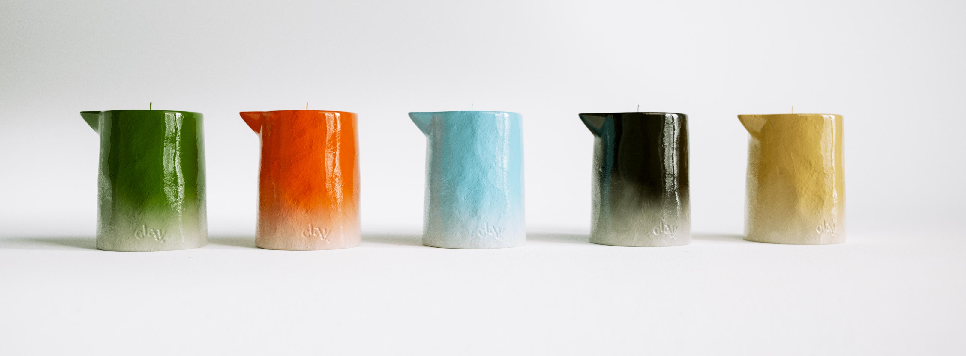 A tight lineup of five CLAY pitcher-form candle samples side by side against a plain white ground, each showing the brand's signature ombré lacquer fading into a pale raw base stamped with the lowercase "clay." deboss. Left to right the row runs deep green, burnt orange, pale blue, near-black charcoal and warm tan, the first three matching the confirmed Champagne, Amber and Vetiver colourways, the black and tan two additional development samples not yet assigned to a released scent. Shown unboxed and unstyled, it's a colour and wax-finish reference rather than a product shot — the kind of side-by-side comparison a multi-SKU colour system gets checked against before a new scent is signed off. Brand identity and colour development for a growing candle range.