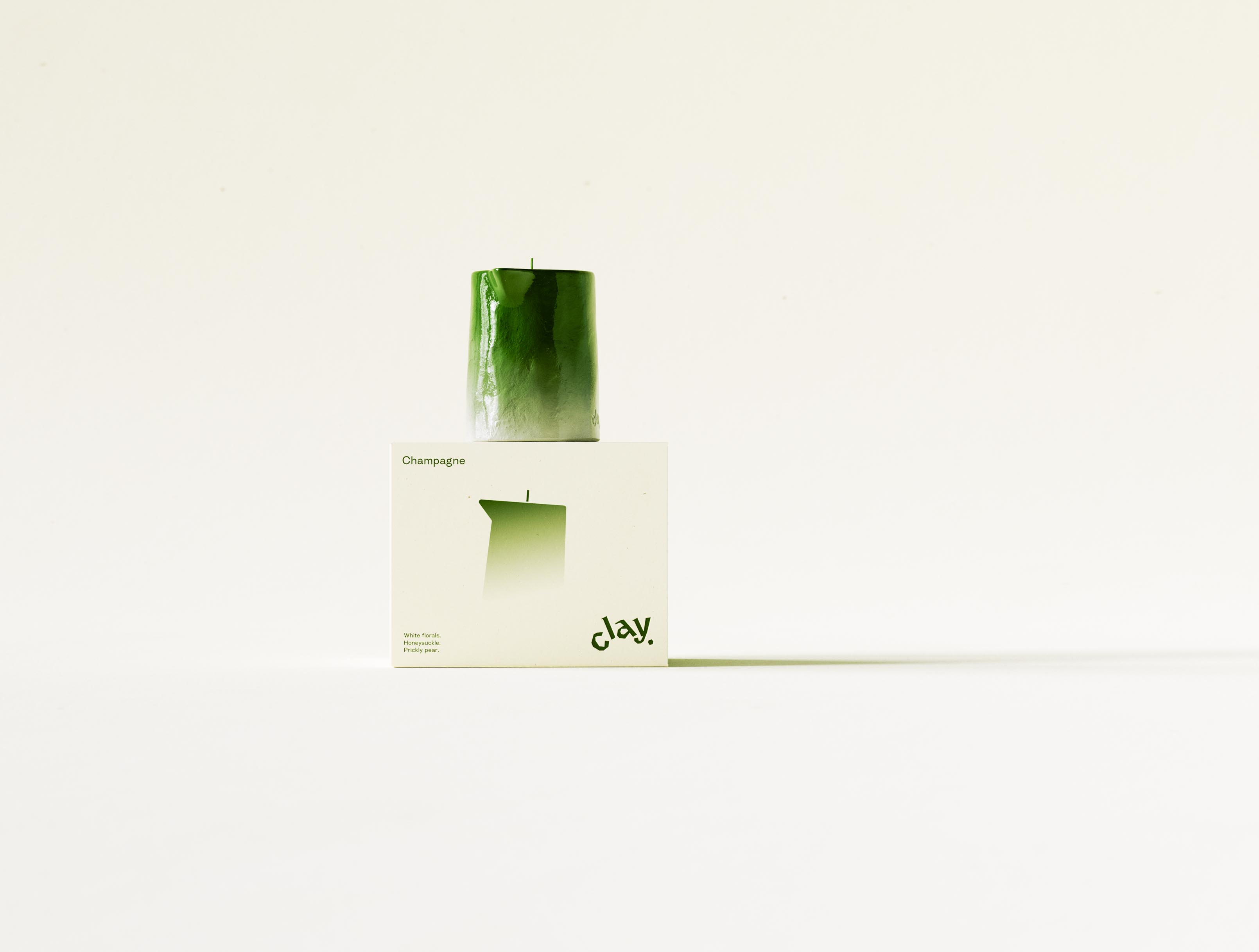 A single CLAY Champagne candle centred on top of its closed carton, both lit softly against a warm cream sweep so the glossy green pitcher form and the printed front face read as one still life. The candle's ombré lacquer runs from saturated green at the pinched spout to a pale raw base stamped with the lowercase "clay." deboss, while the carton beneath repeats the scent name, the gradient candle silhouette, the scent notes and the olive "clay." wordmark. Held dead-centre with generous negative space either side, the frame favours quiet product theatre over busy composition. Bringing a new candle collection to launch — the object, the printed system and the photography all resolved to one calm image — is packaging and art direction AOFP Studio carries through from concept to final shot.