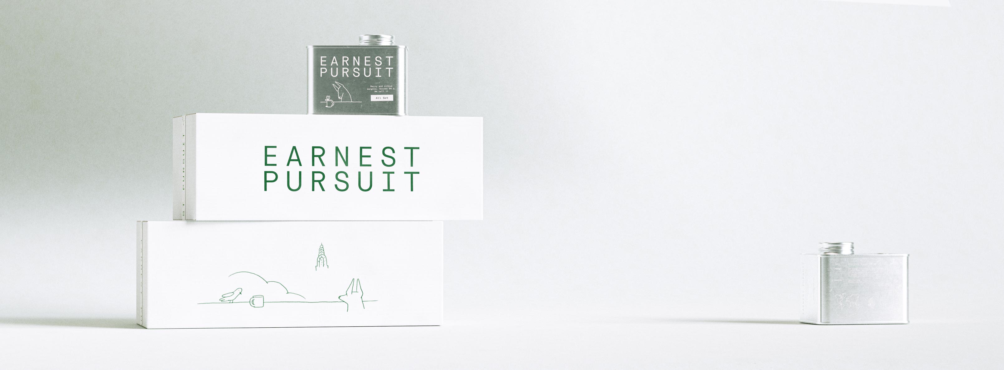 Two long white shipping boxes lie stacked horizontally against a gentle grey gradient, the upper carton carrying the forest-green EARNEST PURSUIT wordmark and the lower one the delicate single-line skyline illustration of a bird, cup, cloud and city spire. A forest-green tin sits on top, its label bearing a small drawing and an "All Day" descriptor, while a bare silver tin rests on the surface to the right. The cartons are matte uncoated corrugate printed in a single green ink; the tin is lithographed metal. The photograph highlights structural packaging design, a coordinated tin-and-carton system and print production — a shipping-packaging programme with matched illustration and typography by Art of Packaging.