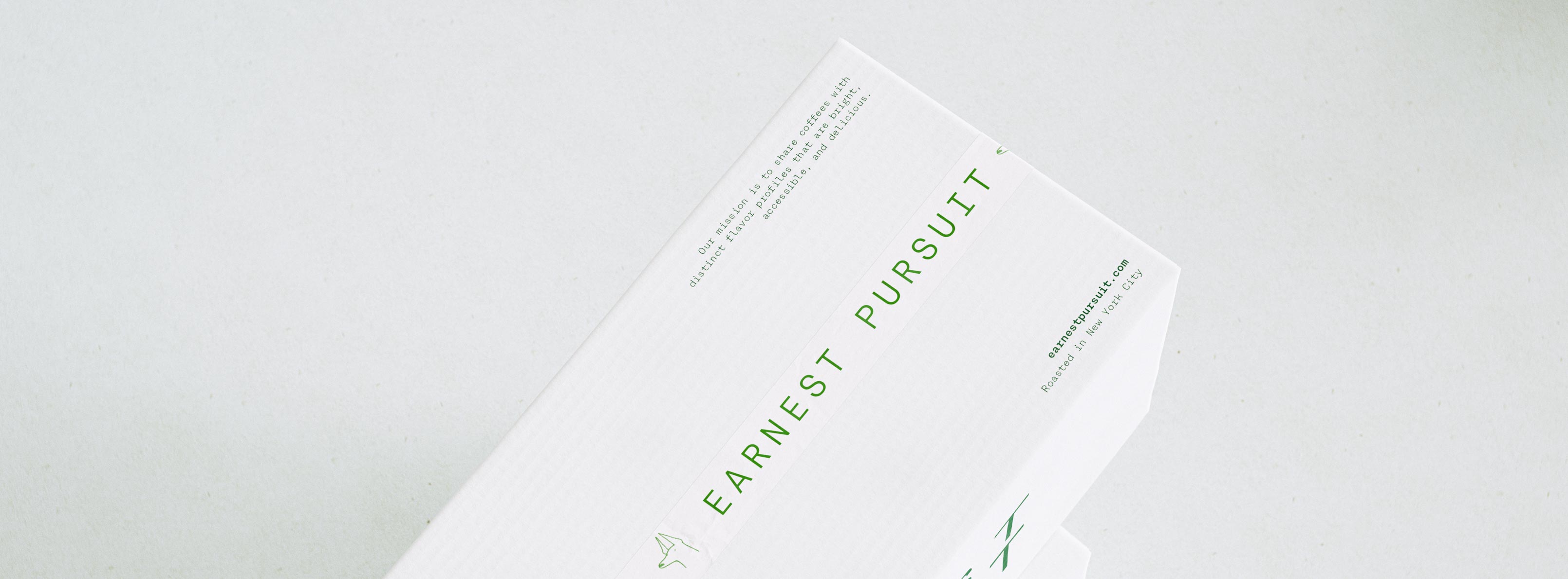 A macro of the top edge of a white Earnest Pursuit mailer, angled so the letter-spaced green EARNEST PURSUIT wordmark runs along the box beside its small-print mission statement: "Our mission is to share coffees with distinct flavor profiles that are bright, accessible, and delicious." Below sit the earnestpursuit.com URL and the "Roasted in New York City" line, with a small skyline glyph tucked in. The carton is uncoated corrugate printed in a single forest-green ink, the copy set in a fine, monospaced-feeling grotesque. The frame foregrounds information design and typographic hierarchy — brand identity and packaging for a new coffee launch from a New York roaster, art direction by AOFP Studio.
