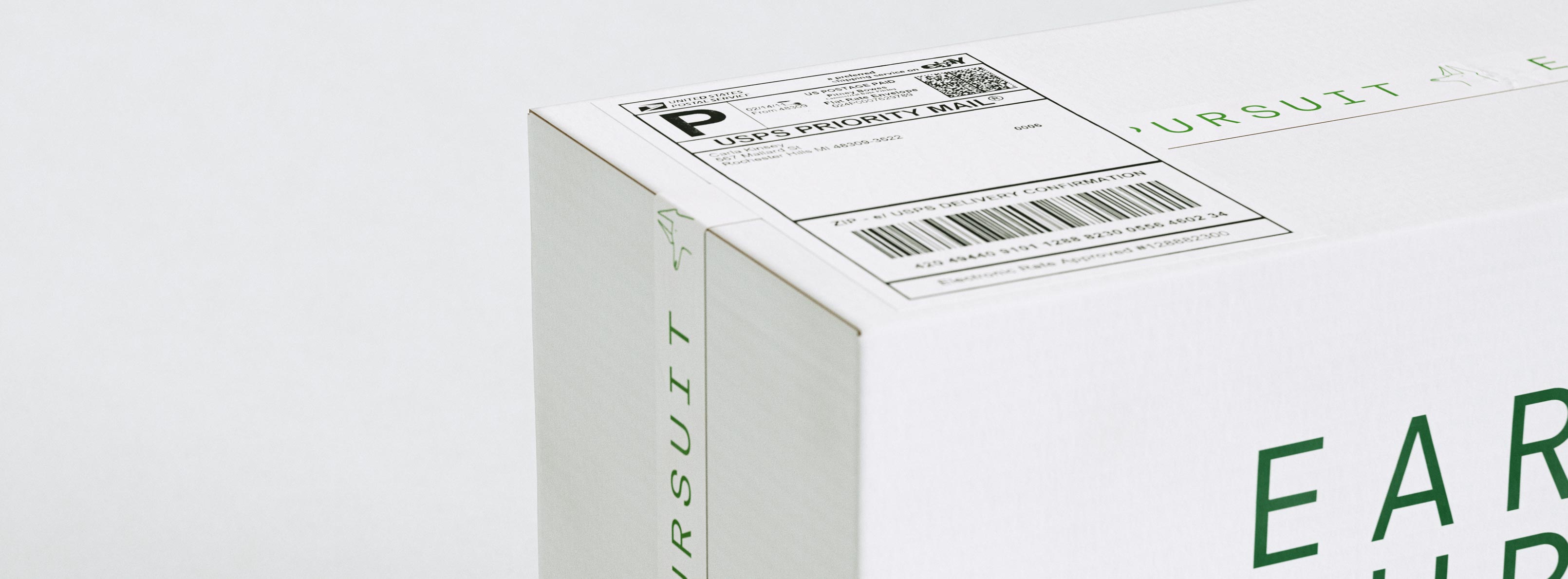 An extreme close-up of the corner of a white shipping mailer, shot at a low angle so the box edge cuts diagonally across the frame. A USPS Priority Mail label sits on the top face, its barcode and "P" mark crisp against the matte board, while the forest-green EARNEST PURSUIT wordmark and a tiny single-line city glyph run along the visible side panel. The carton is uncoated corrugate with restrained one-colour green printing and generous negative space. This macro highlights the finish and detailing of the shipper — the typography, label placement and material feel — the sort of packaging design for specialty-coffee brands that Art of Packaging develops.