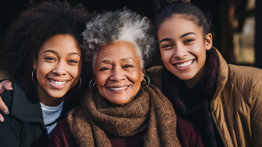 Smiling elderly woman with gray curly hair and two younger women embracing her on each side.