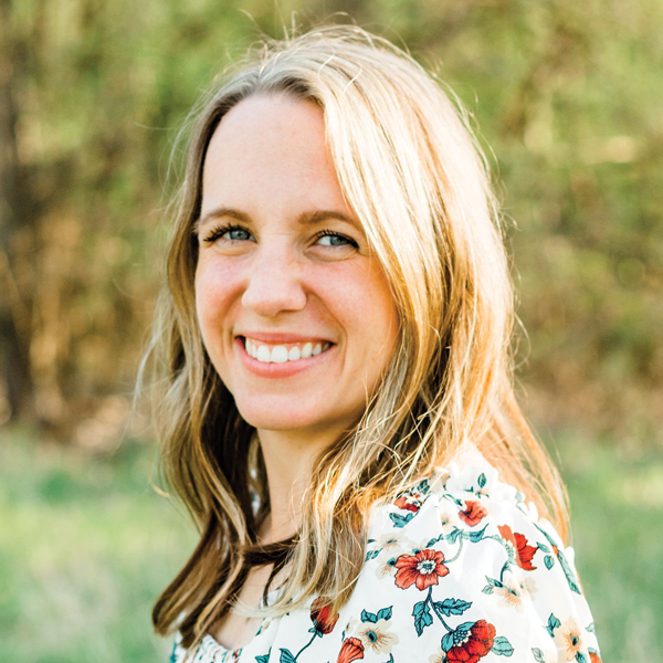 Smiling woman with long blonde hair wearing a white floral blouse standing outdoors with blurred greenery background.