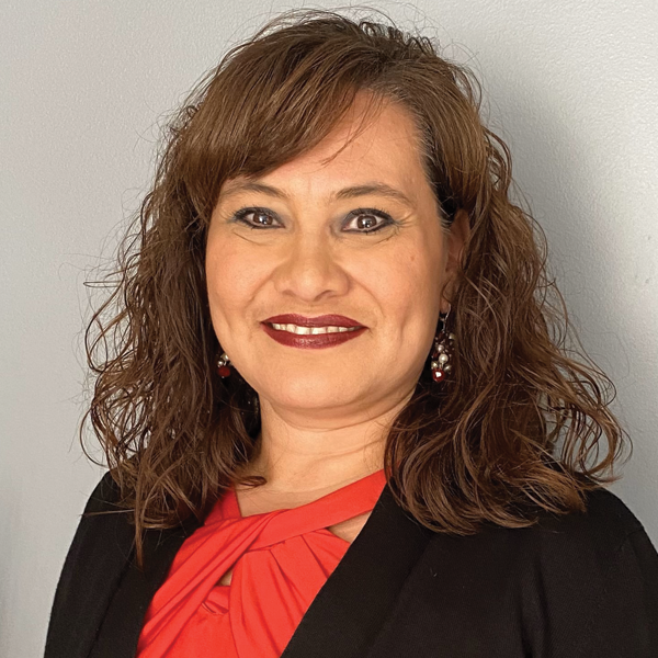 Smiling woman with curly brown hair wearing red top and black blazer against a plain light gray background.