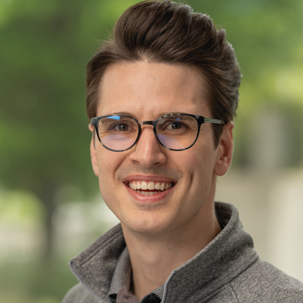 Smiling young man with glasses and styled brown hair wearing a gray fleece jacket outdoors.