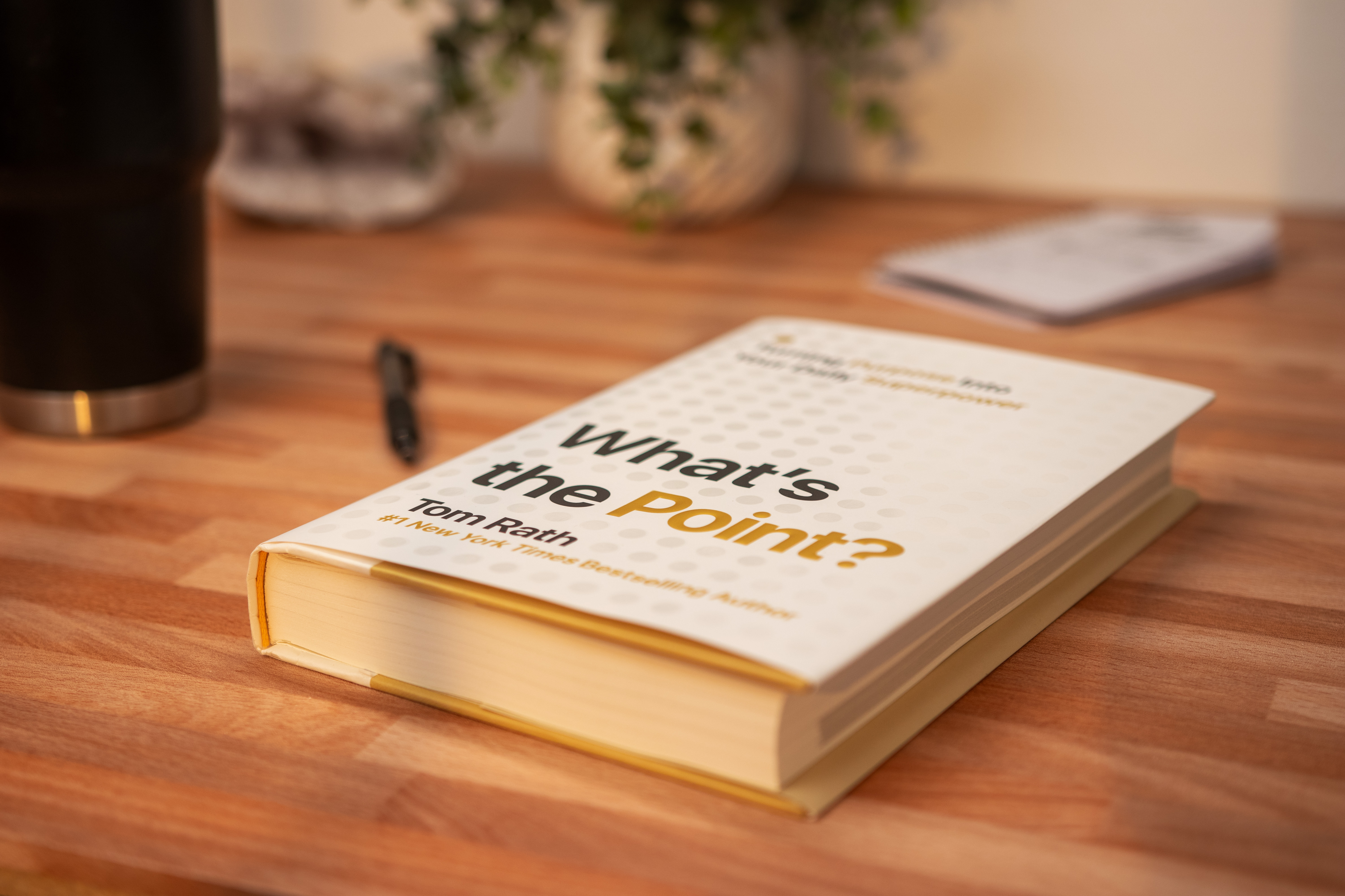 A book titled What's the Point? by Tom Rath lays on a wood desk with a coffee mug and pen in the bakground.