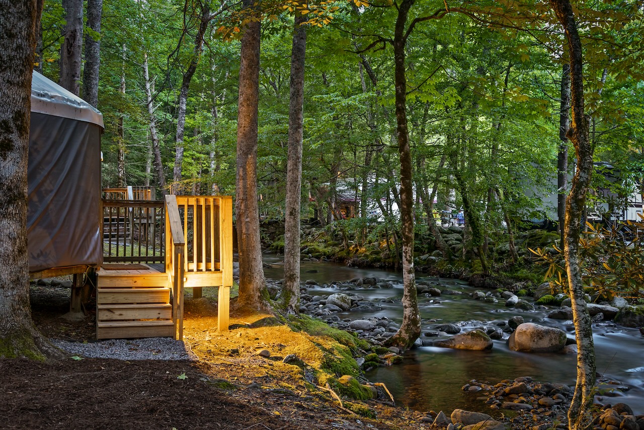 Nighttime view of a Roamstead yurt and illuminated wooden walkway near a creek.