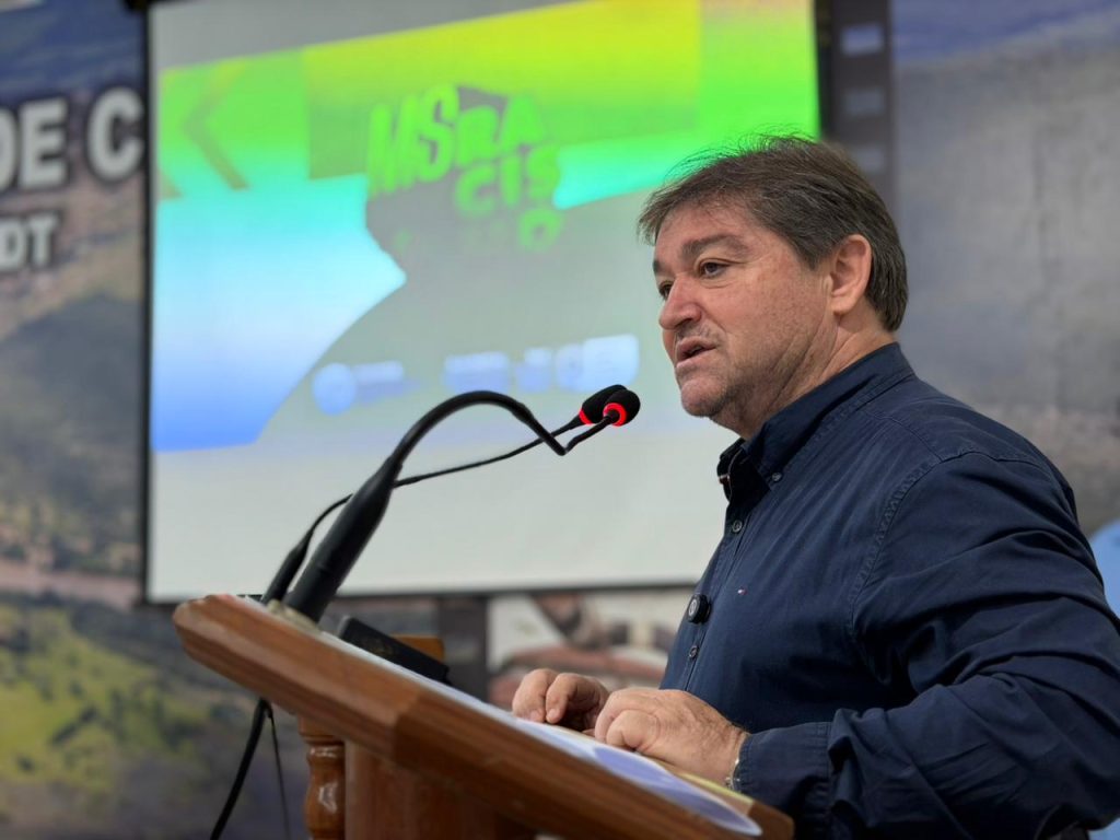Um homem branco, vestindo uma camisa social azul escura, no palco. Ele está falando em um microfone com expressão séria, durante o evento.