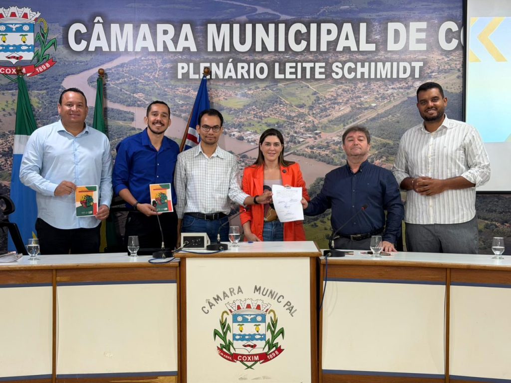 Foto de grupo no plenário, com seis pessoas (quatro homens e uma mulher de blazer laranja, e outro homem) em pé atrás de uma mesa, posando para a foto após a assinatura. A mulher no centro segura um documento assinado, e o fundo é um painel panorâmico da cidade de Coxim