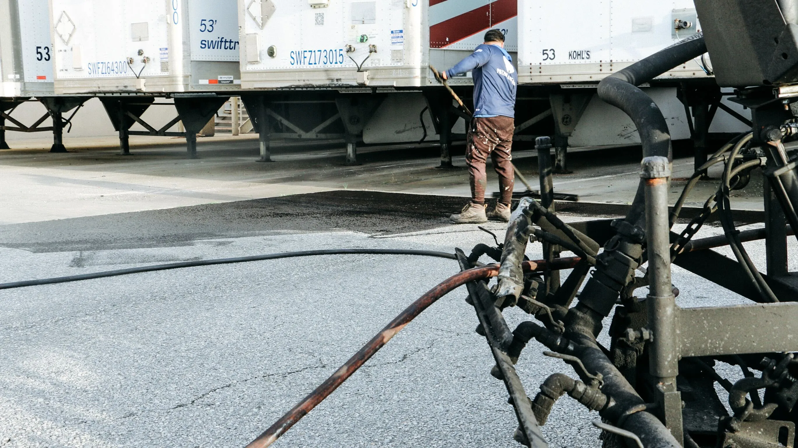 A man in a blue shirt and brown pants is cleaning a road with a hose connected to a truck, while a large truck with a red and white color scheme is parked nearby, and a tree is visible in the background.