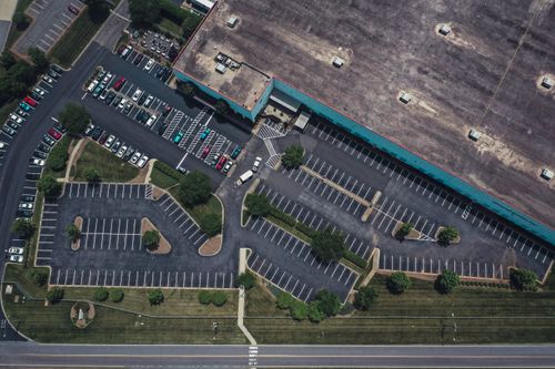 Aerial view of a large parking lot next to a building with some cars parked and green areas.