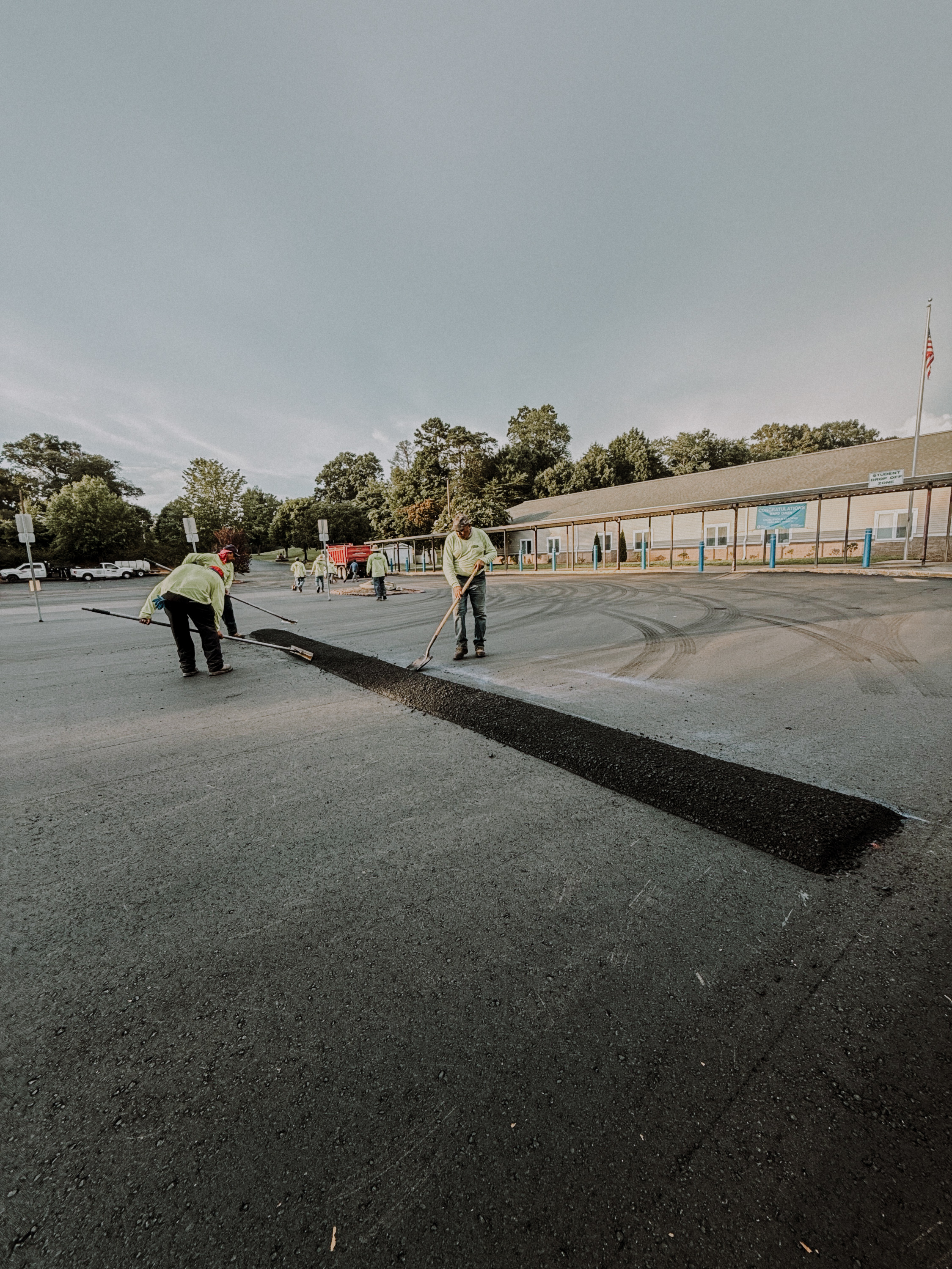 Workers in yellow shirts spreading black asphalt on a parking lot near a school building.