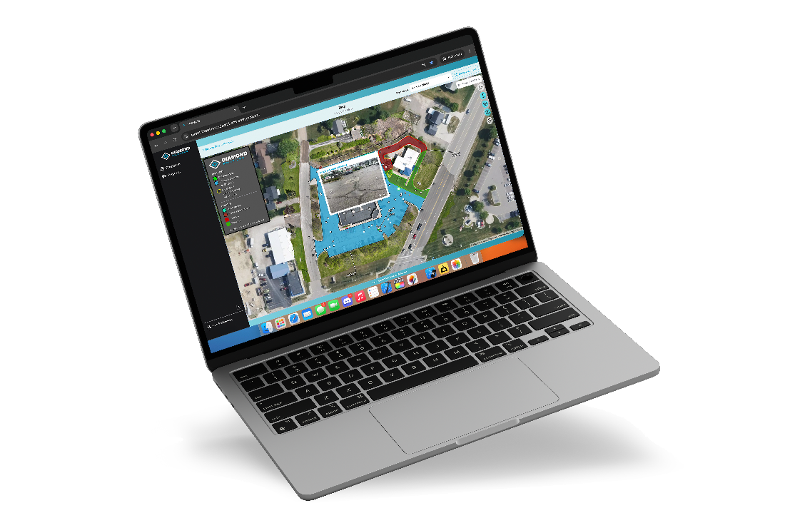Modern silver laptop tilted at an angle showing a map-marking software on its screen with satellite view highlighting buildings and roads using blue, red, and green boundary overlays.
