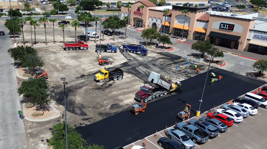 Construction workers paving a large parking lot in front of a shopping plaza with stores including Petco, 5.11 Tactical, and OshKosh B'gosh, with trucks and equipment spreading asphalt and rolling it flat.