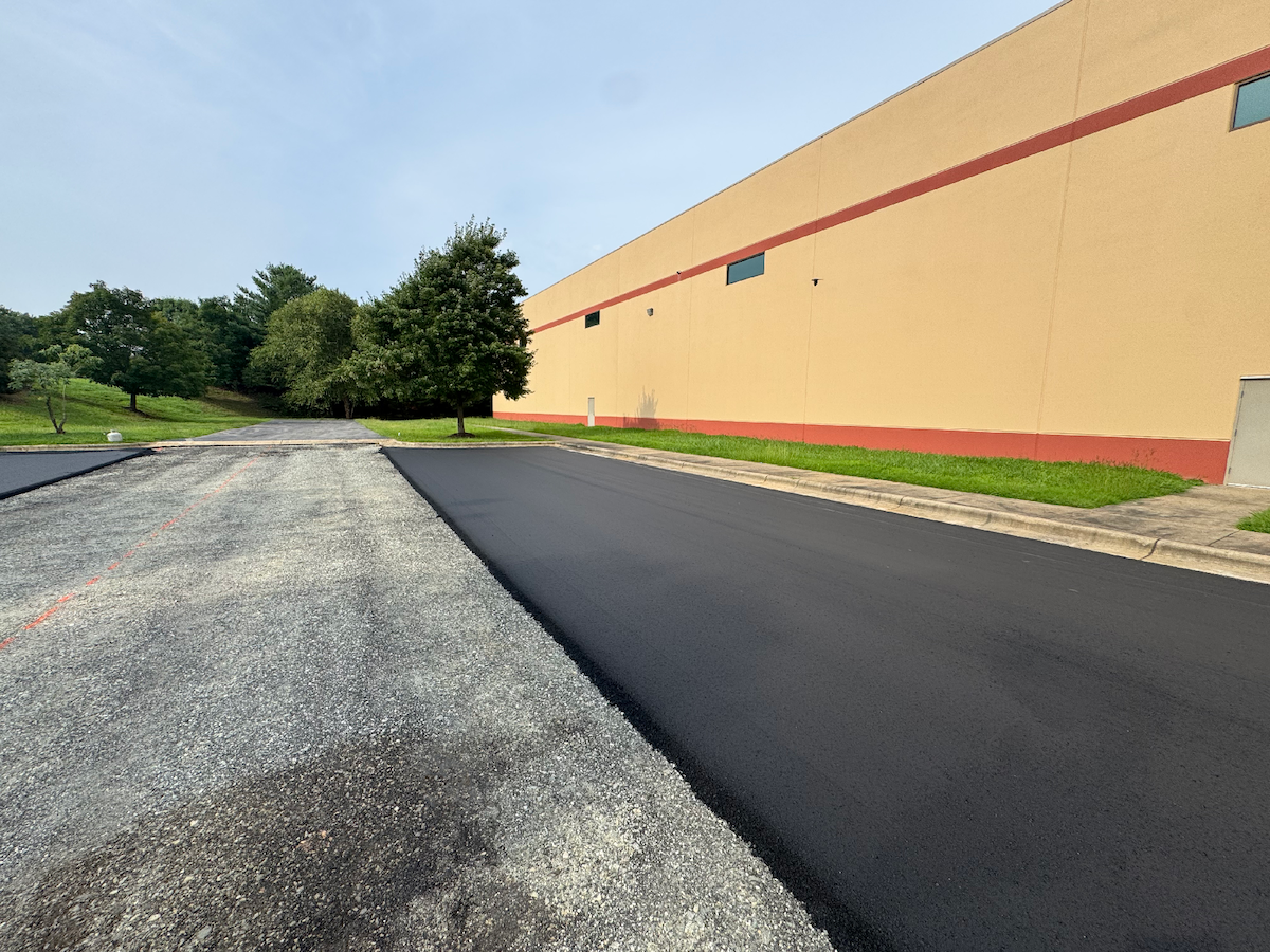 Construction workers paving a large parking lot in front of a shopping plaza with stores including Petco, 5.11 Tactical, and OshKosh B'gosh, with trucks and equipment spreading asphalt and rolling it flat.