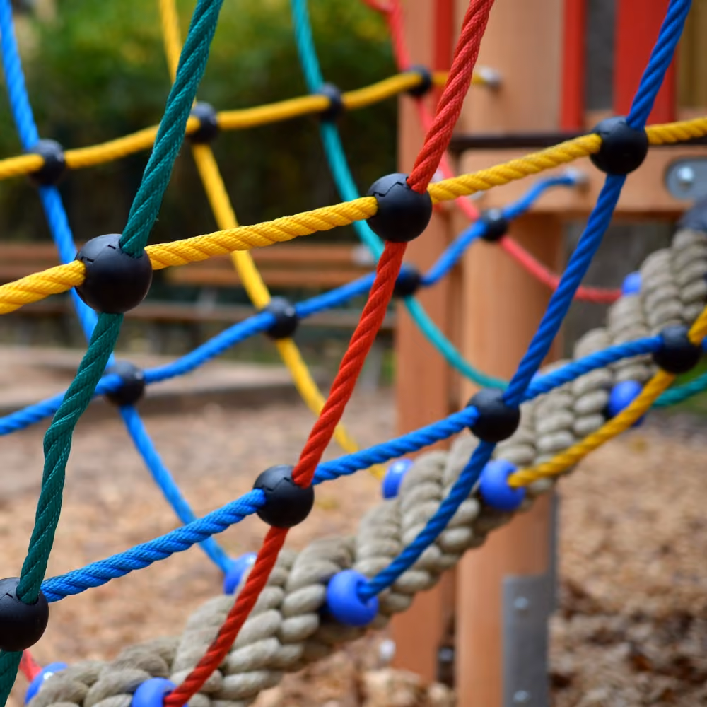 Mehrfarbiges Kletternetz mit roten, grünen, blauen und gelben Seilen auf einem Spielplatz mit Holzelementen und Holzhackschnitzelboden.