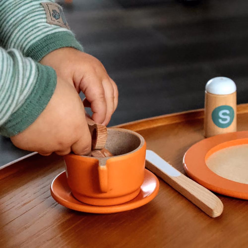 Kinderhände spielen mit einem kleinen Holzlöffel in einer orangefarbenen Schale auf einem Holztisch, daneben ein hölzernes Messer, ein Salzerstreuer mit grünem Aufkleber und ein Teller mit orangefarbenem Rand.