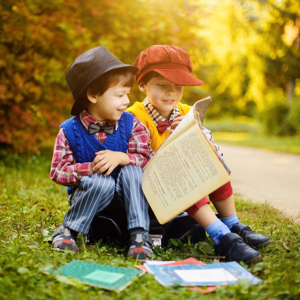 Zwei Jungen sitzen auf dem Gras im Freien, die Sonne scheint, einer liest ein Buch, der andere lächelt und hört zu, beide tragen bunte Westen, Hemden und Hüte.