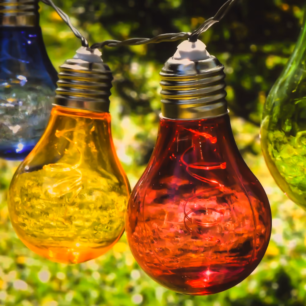 Nahaufnahme von bunten Glühbirnenlichtern in Rot, Gelb, Blau und Grün, die an einem Kabel hängen, mit einem unscharfen grünen Hintergrund.